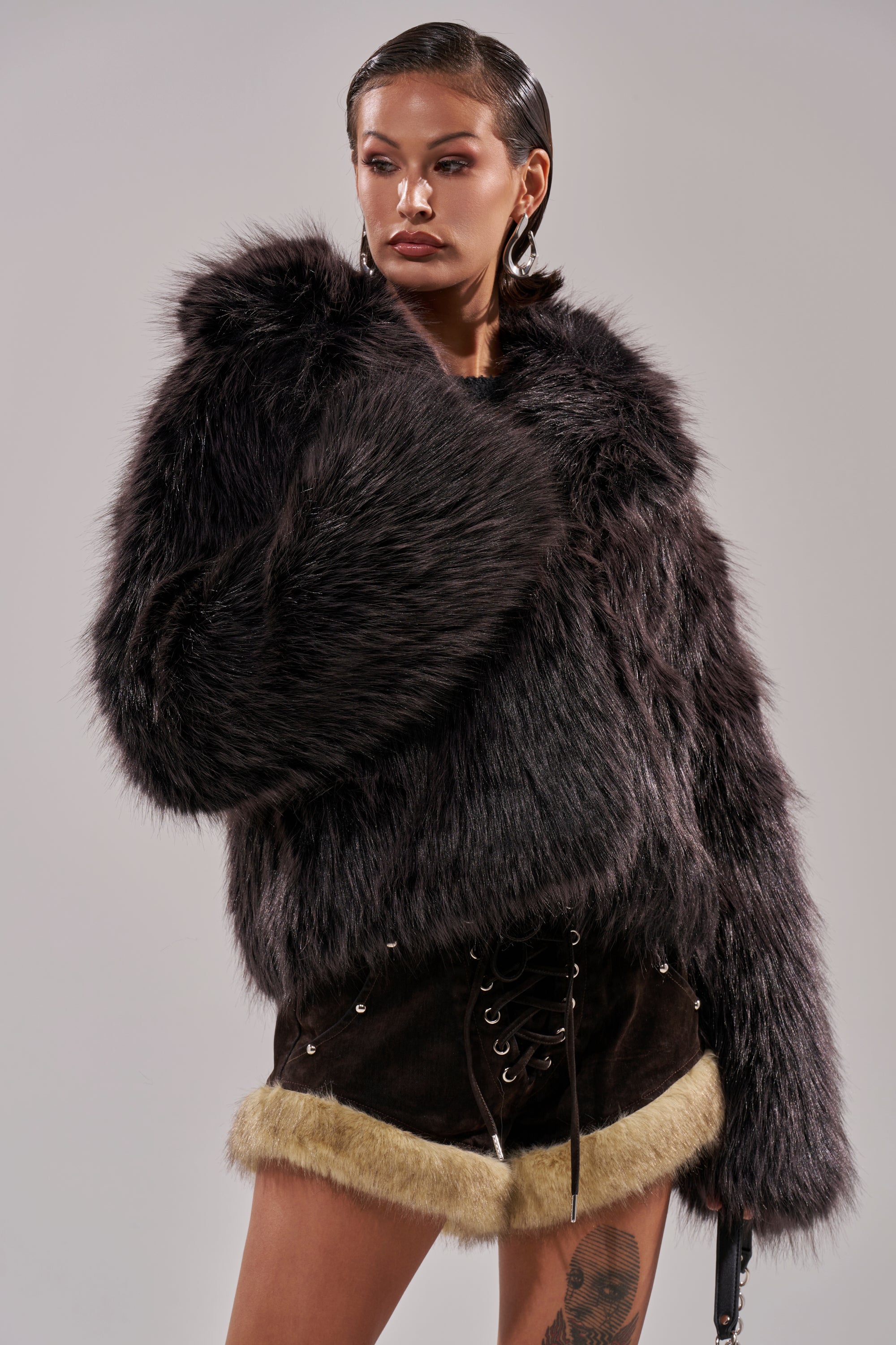 A person stands confidently in the DUSK TIL DAWN FAUX FUR COAT, styled with black lace-up shorts featuring beige fur trim and hoop earrings, against a plain light background. 4