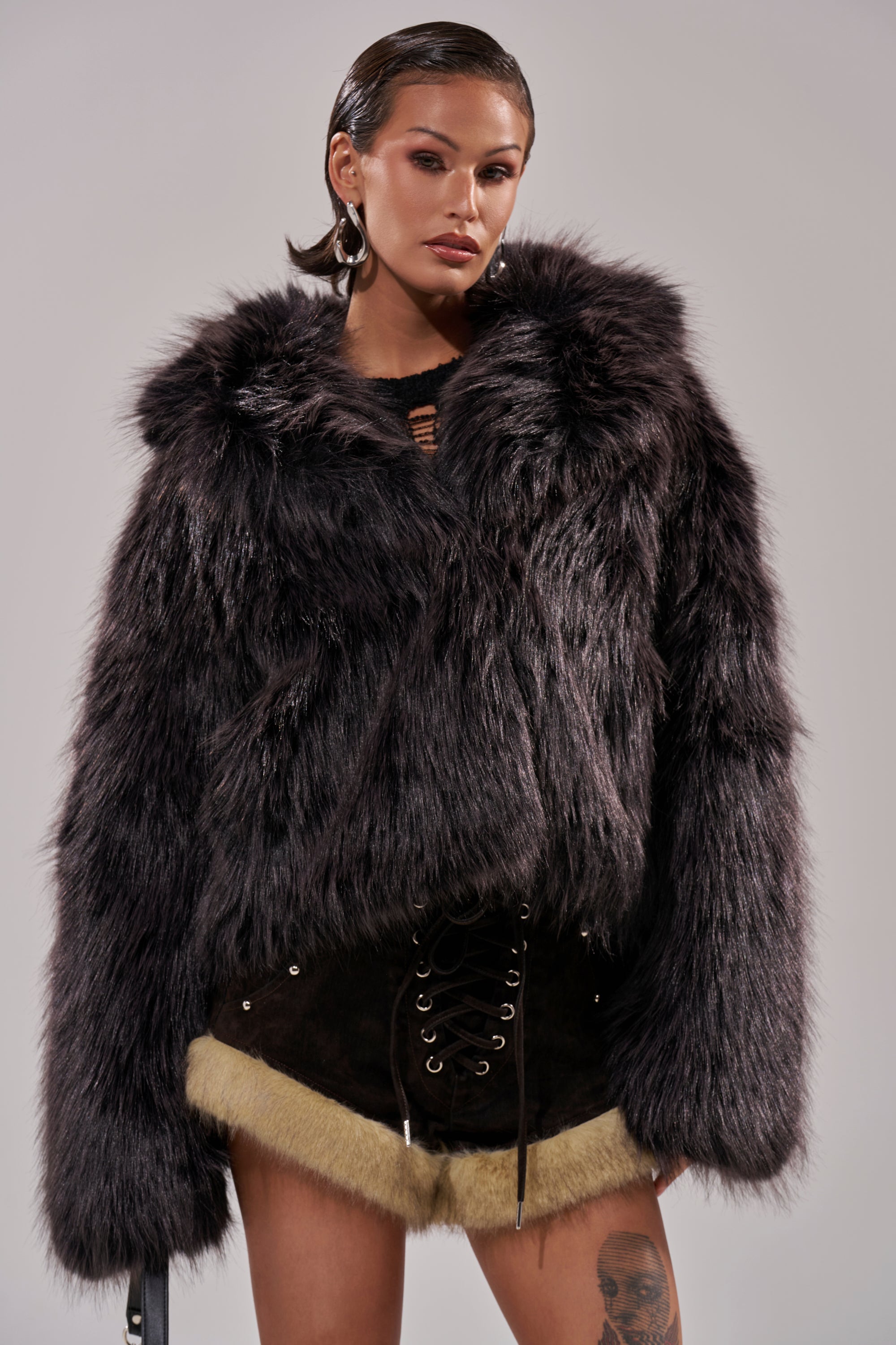 A woman in the luxurious DUSK TIL DAWN FAUX FUR COAT and black lace-up shorts with beige trim poses against a gray background. She has slicked-back hair, bold makeup, hoop earrings, and a visible leg tattoo. 5