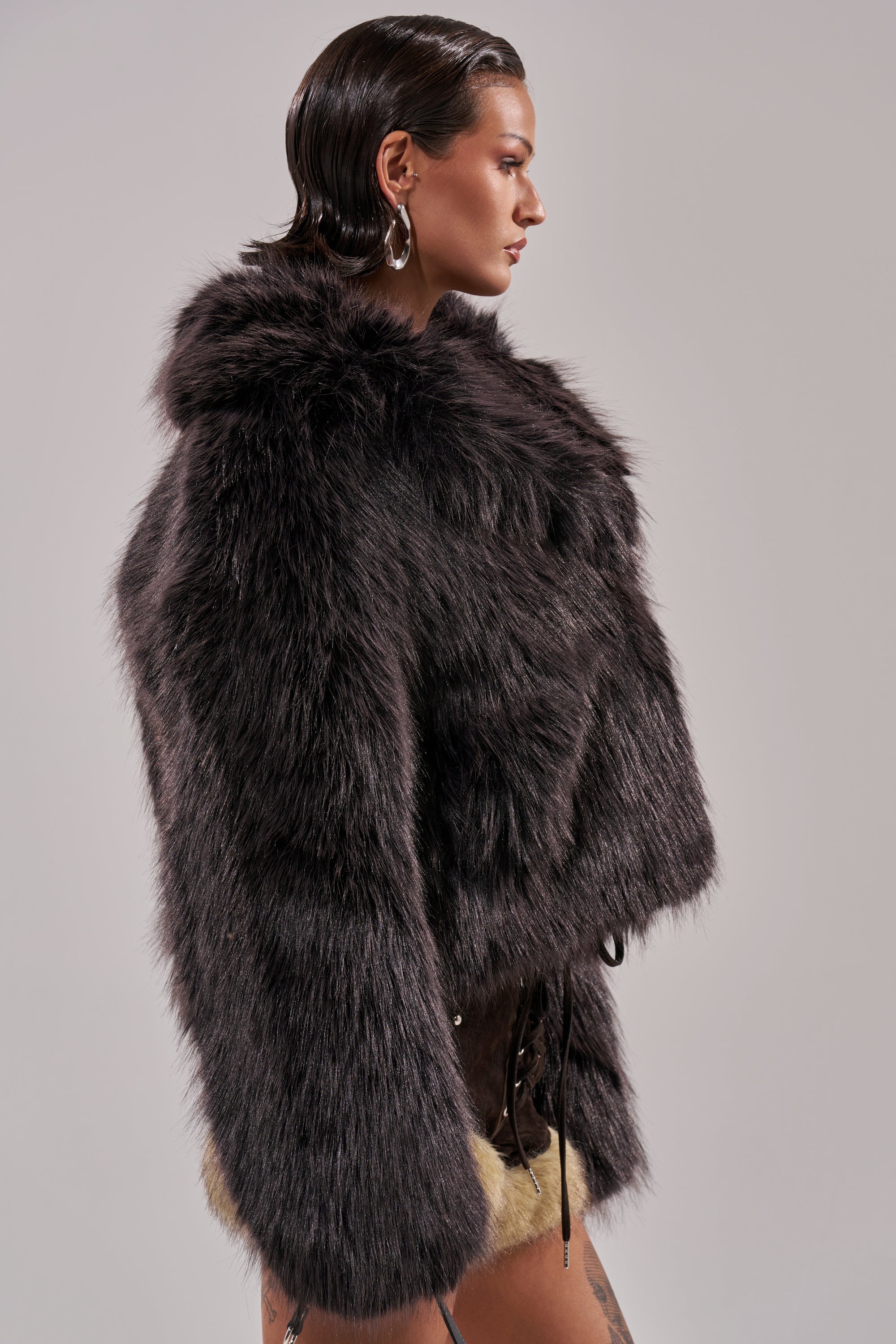 A person with slicked-back hair and hoop earrings models the DUSK TIL DAWN FAUX FUR COAT and cream shorts, standing in profile against a plain light background. 6