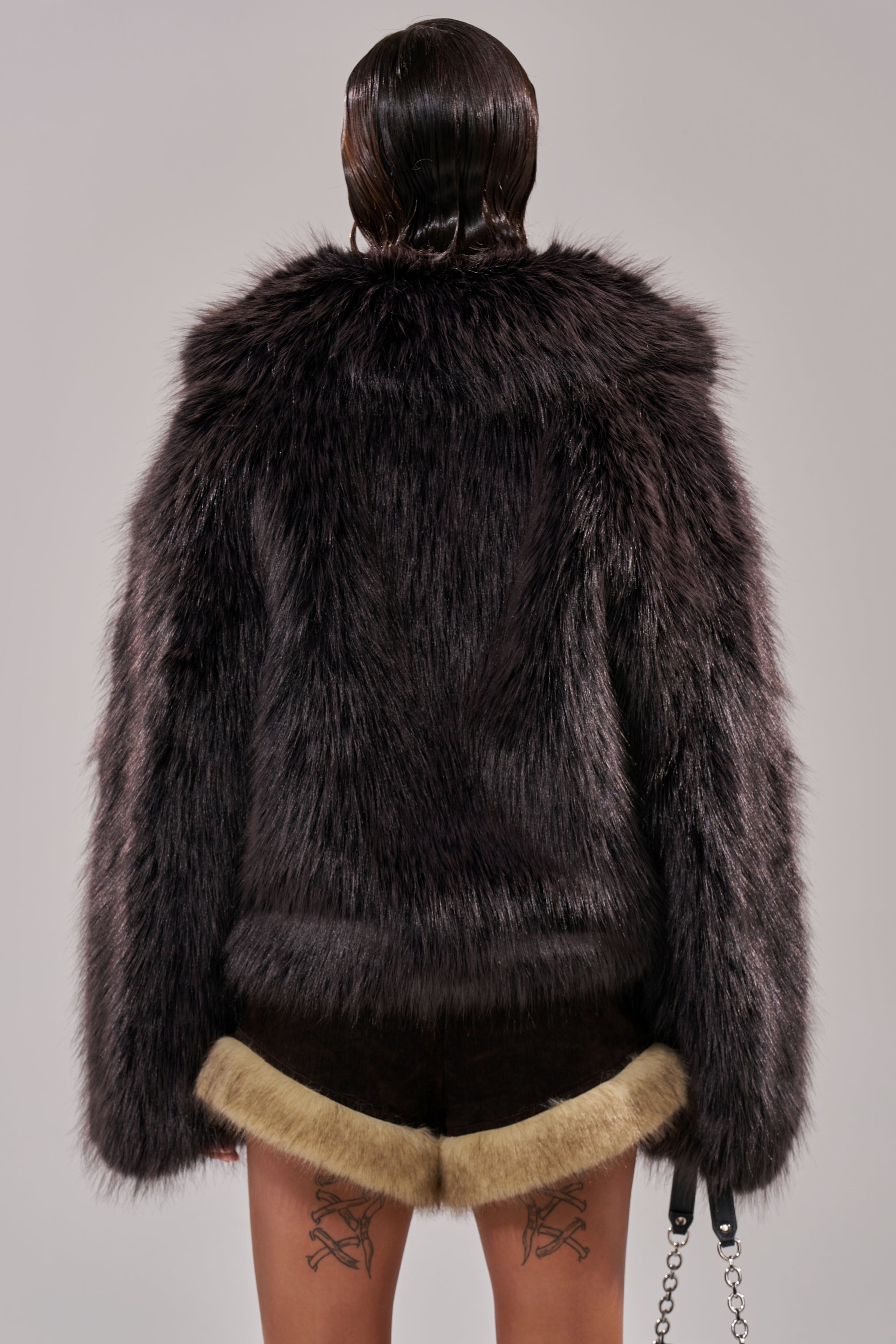 A person with slicked-back dark hair is seen from behind in the oversized DUSK TIL DAWN FAUX FUR COAT and dark shorts with light fur trim. Crossed sword tattoos on the backs of their thighs complete this bold ensemble. 7