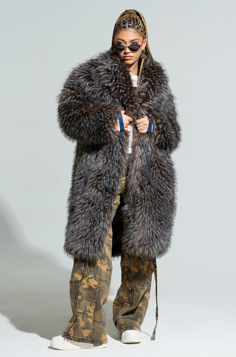 A person wearing sunglasses confidently models the "KEEP TO MYSELF FAUX FUR JACKET," a long and chicly shaggy piece, over a white top and camouflage pants. Their braids are styled neatly as they hold the jacket with a firm grip against a plain light-colored background.
