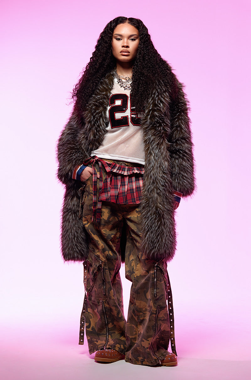 A person with long curly hair wears the chic KEEP TO MYSELF FAUX FUR JACKET over a sports jersey, a plaid shirt tied at the waist, and camouflage pants with straps. They stand against a pink gradient background, exuding a bold, eclectic style.