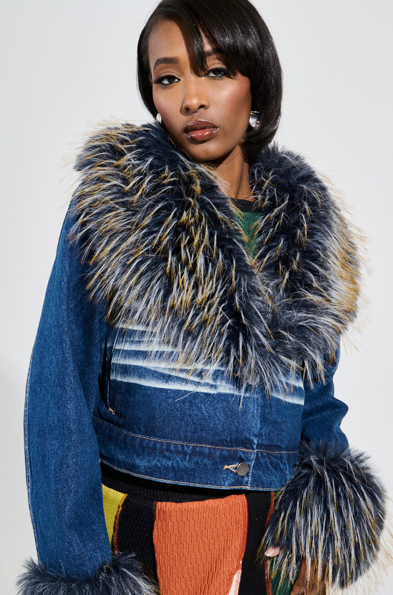 A person wearing the AD ASTRA FUR TRIM JACKET IN DARK BLUE DENIM, featuring a large faux fur collar and cuffs with shades of blue and brown, stands against a plain background, complementing their colorful undergarment.