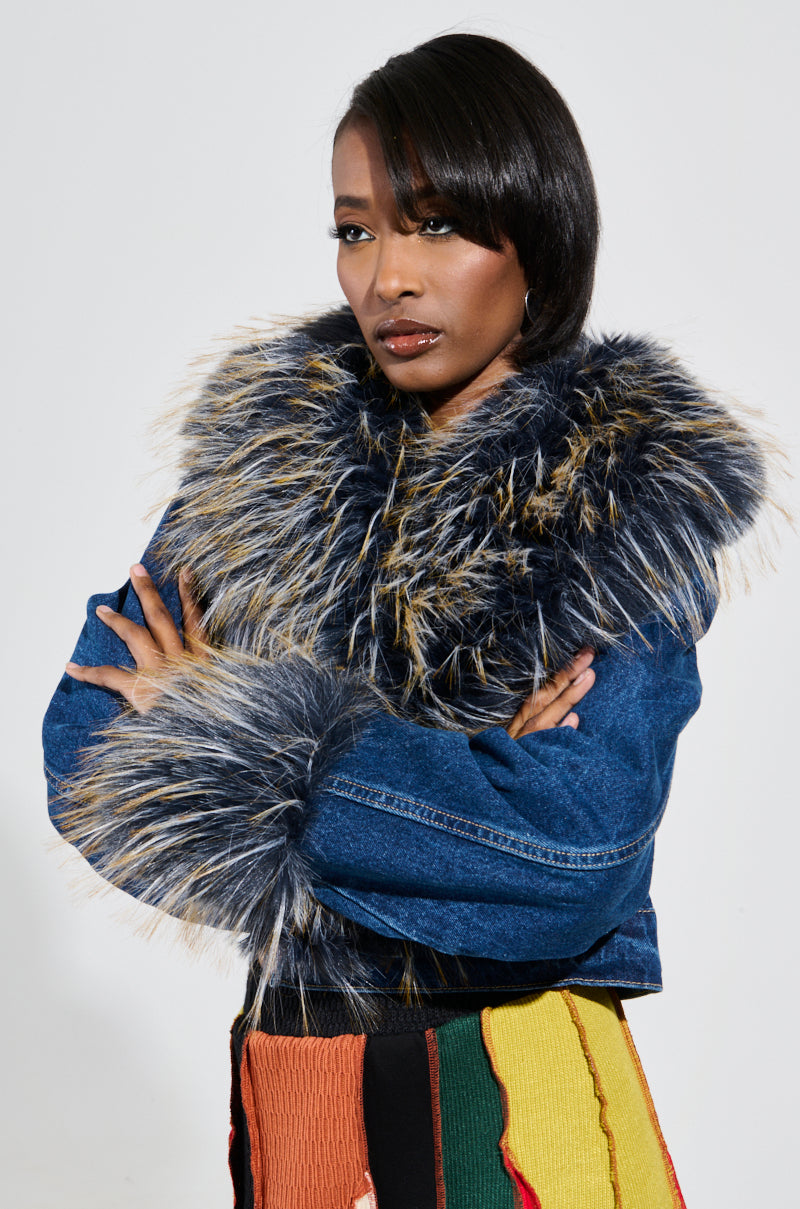A person stands confidently with crossed arms, wearing the AD ASTRA FUR TRIM JACKET IN DARK BLUE DENIM, which features a large fur-trimmed collar and matching furry cuffs. They wear a multicolored skirt and have a sleek hairstyle.