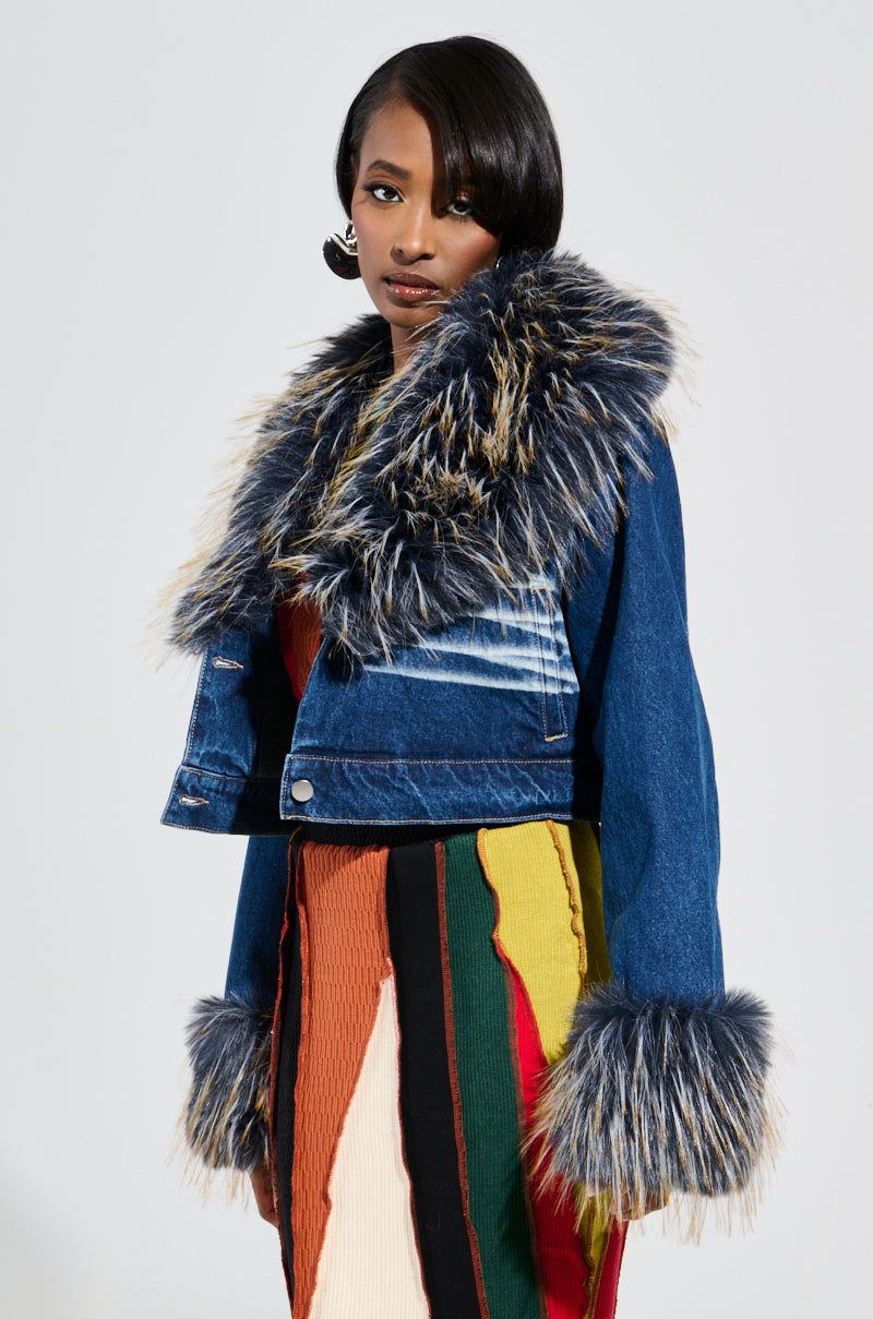 A woman in a chic ensemble poses confidently, wearing the AD ASTRA FUR TRIM JACKET IN DARK BLUE DENIM with fur trim on the collar and cuffs, elegantly paired with a colorful, textured skirt. Her sleek shoulder-length hair complements her serious expression perfectly.