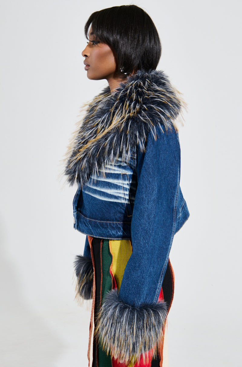 A person in profile wears the AD ASTRA FUR TRIM JACKET IN DARK BLUE DENIM, featuring a large, fluffy fur trim on the collar and cuffs. They have a short bob hairstyle and elegantly pair the jacket with a colorful, striped garment. The background is plain white.