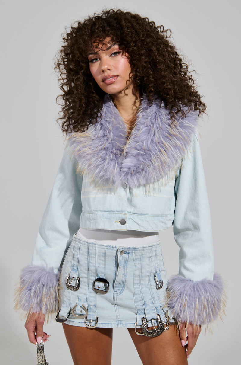 Sporting the AD ASTRA Fur Trim Jacket in light blue denim, a woman with curly hair channels Bratz-inspired fashion. The jacket's purple faux fur accents enhance her stylish look, while the accompanying skirt's metallic buckles and safety pins introduce an edgy flair. She completes her ensemble with a small chain strap purse. 0