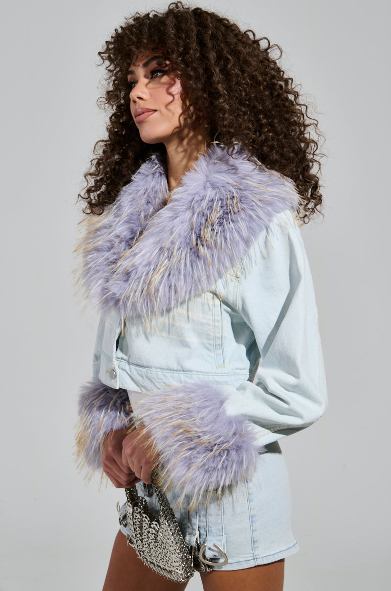 Wearing the "AD ASTRA FUR TRIM JACKET IN LIGHT BLUE DENIM," a woman with curly hair is adorned with lavender faux fur on the collar and cuffs. She holds a small silver handbag and stands in profile against a plain background, channeling Bratz-inspired fashion. 2