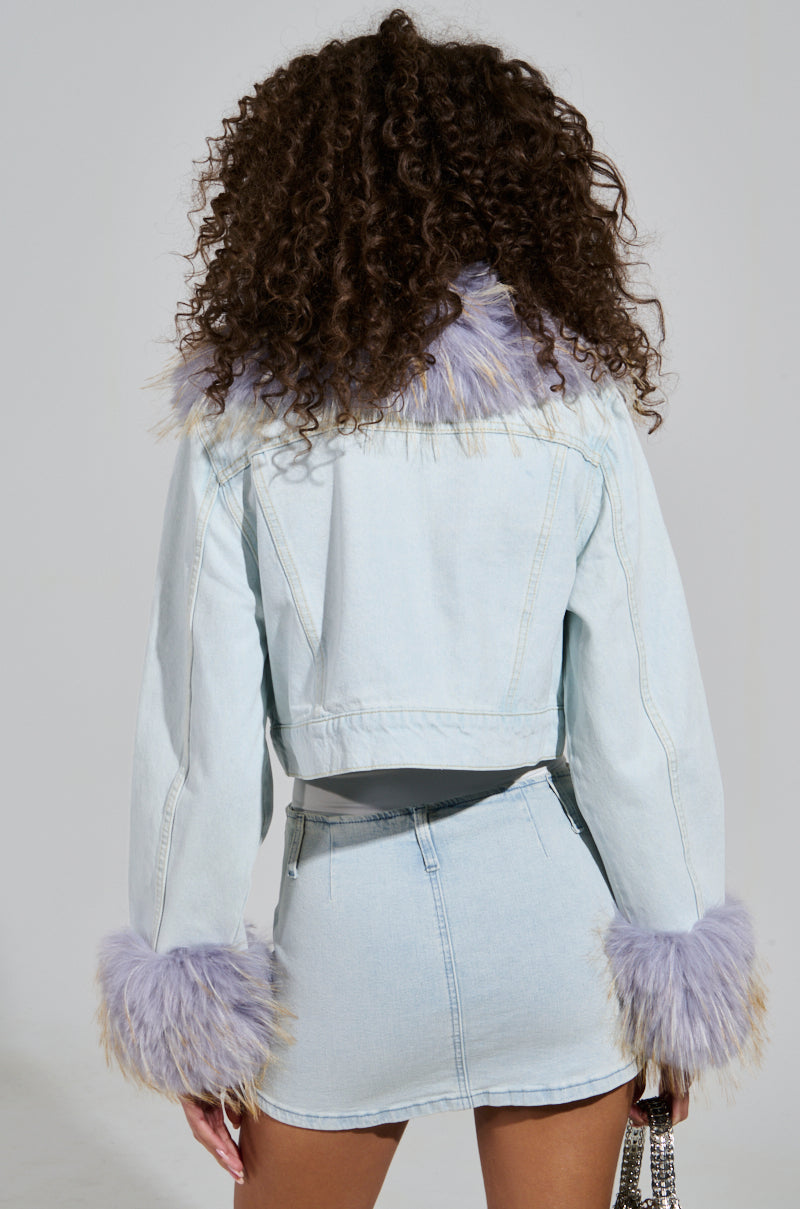 A person with curly hair channels Bratz fashion in the AD ASTRA FUR TRIM JACKET IN LIGHT BLUE DENIM and a matching skirt. The jacket, adorned with a purple fur trim on the collar and cuffs, pairs perfectly with their small, sparkly handbag. They turn away from the camera, embodying a playful yet stylish aesthetic. 3