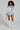 A woman with curly hair captures a Bratz vibe in her AD ASTRA FUR TRIM JACKET IN LIGHT BLUE DENIM, a mini skirt adorned with patches, a white top, knee-high white lace-up boots, and a small silver handbag.
