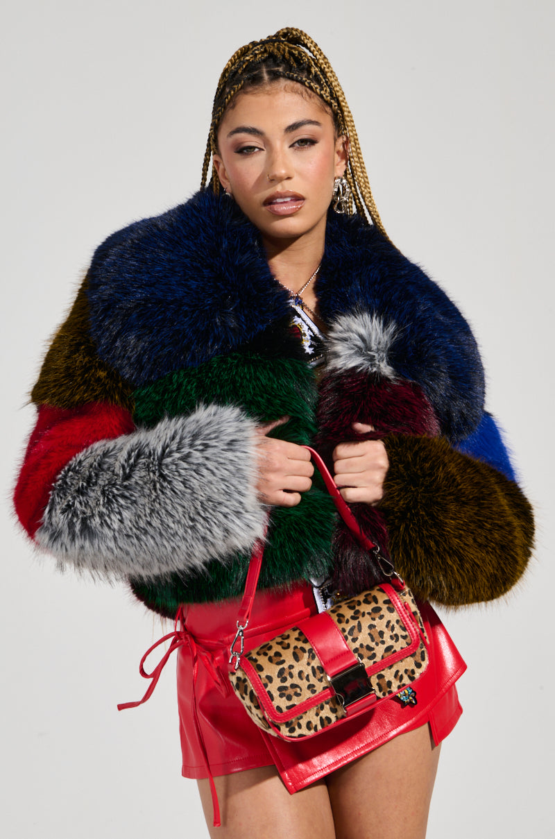 Dressed in the RIOT PATCHWORK FAUX FUR coat, a person with braided hair showcases a vibrant fusion of textures. They pair it elegantly with a red leather skirt and carry a leopard-print handbag, creating an outfit that exudes eclectic charm against the plain background.