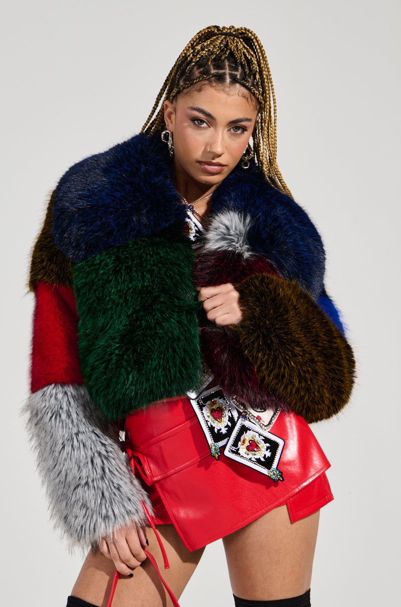 A person with braided hair highlights the vibrant RIOT PATCHWORK FAUX FUR coat, designed in a striking color-block pattern of blue, green, red, brown, and gray. Complemented by a red skirt-like ensemble embellished with card-themed patches, they pose against a simple gray backdrop.