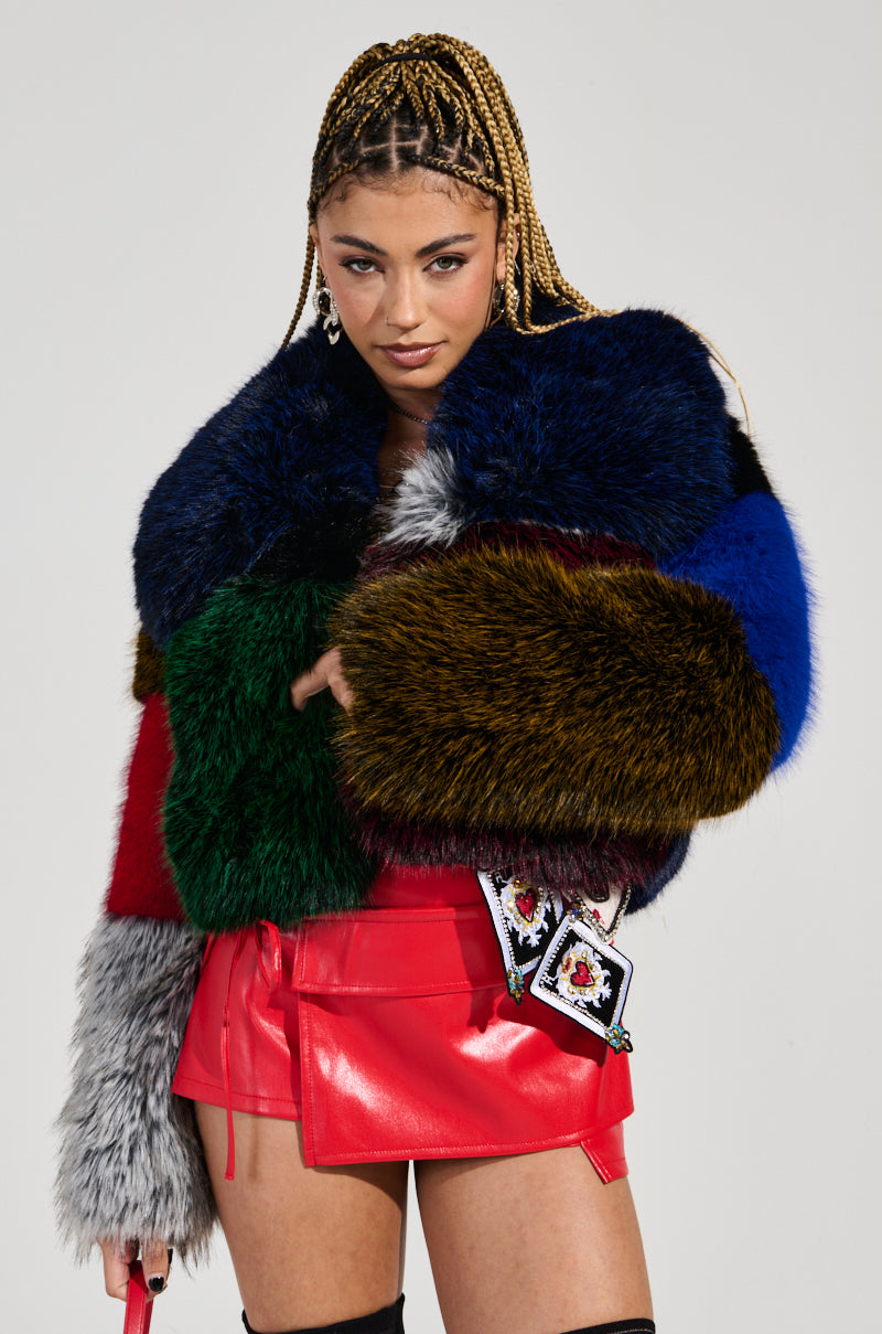 A woman with braided hair exudes style in a RIOT PATCHWORK FAUX FUR jacket paired with a red leather skirt. She holds a small purse embellished with decorative patches, and the plain background accentuates her fashion-forward, color-block design ensemble.