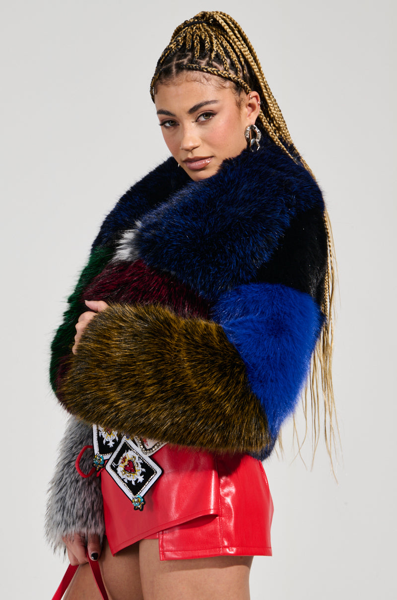 Wearing the RIOT PATCHWORK FAUX FUR jacket, a person with braided hair stands out in a vibrant mix of colors. Their ensemble includes bright red leather pants paired with a coordinating bag adorned with decorative patches, forming an eye-catching color-block design. Posing confidently against a neutral backdrop, they highlight their bold and cropped silhouette.
