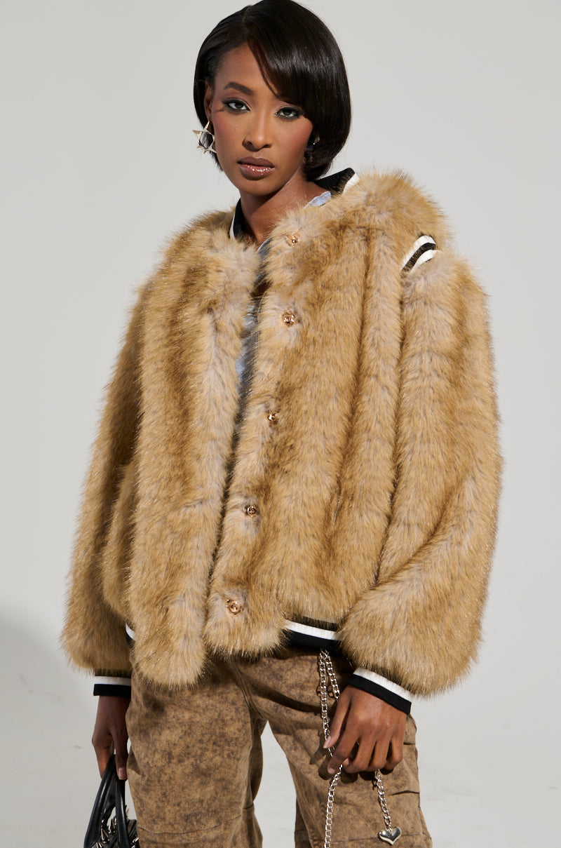 A person wearing the RINGO FUR BOMBER, featuring a brown faux fur design with a ribbed knit collar, paired with brown pants stands confidently against a plain background. They hold a black handbag and have short, styled hair.