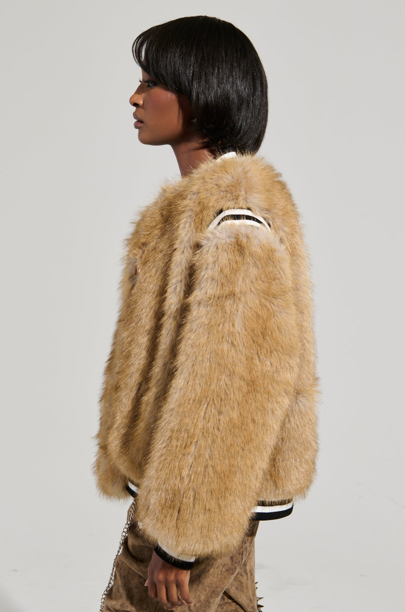 A person wearing the RINGO FUR BOMBER, an oversized faux fur jacket in light brown with black and white striped sleeve accents, stands against a plain background, facing left. The individual has short, dark hair.
