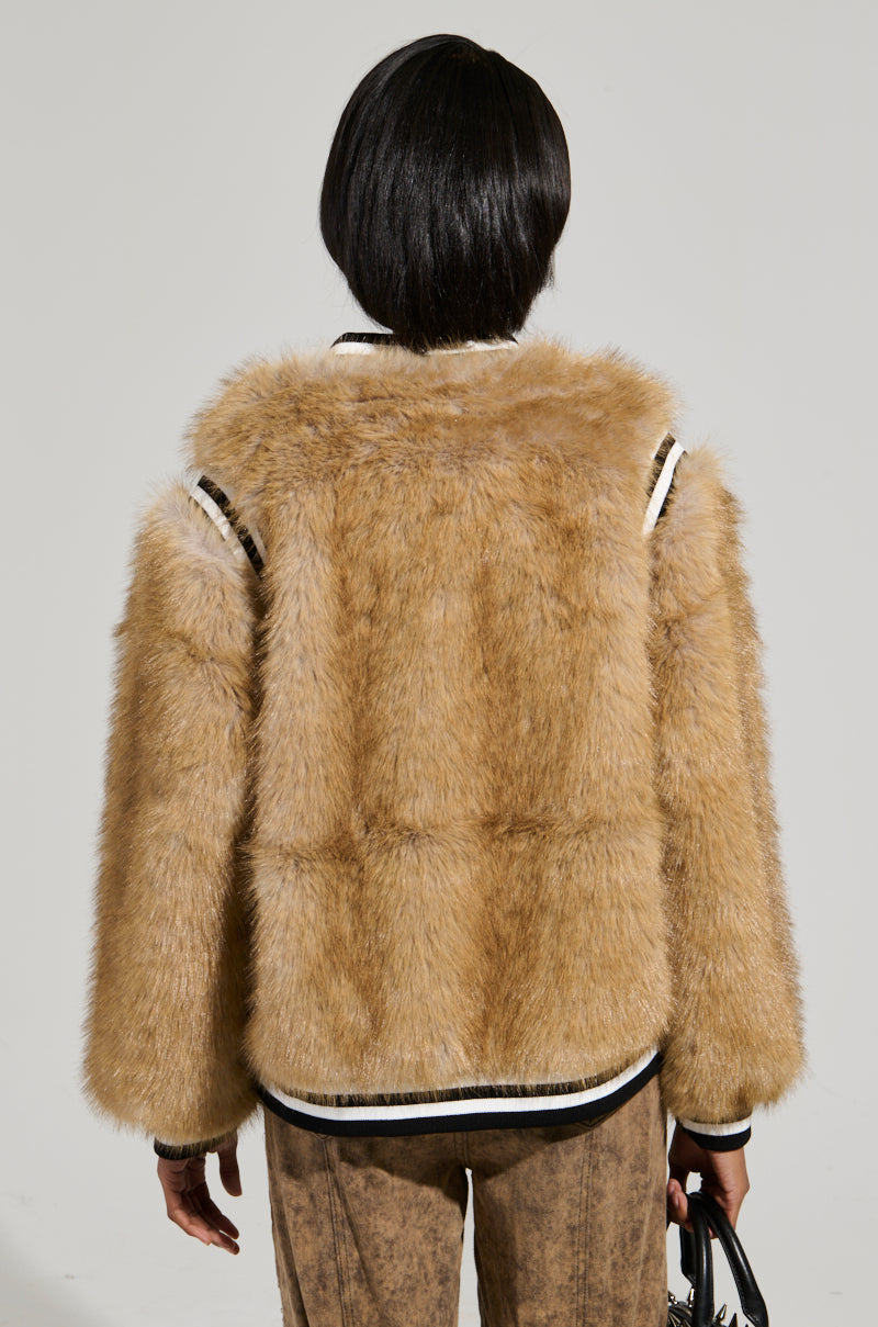 A person with short hair is wearing the RINGO FUR BOMBER, an oversized faux fur jacket with black and white trim, seen from the back while holding a small black handbag against a plain light gray background.