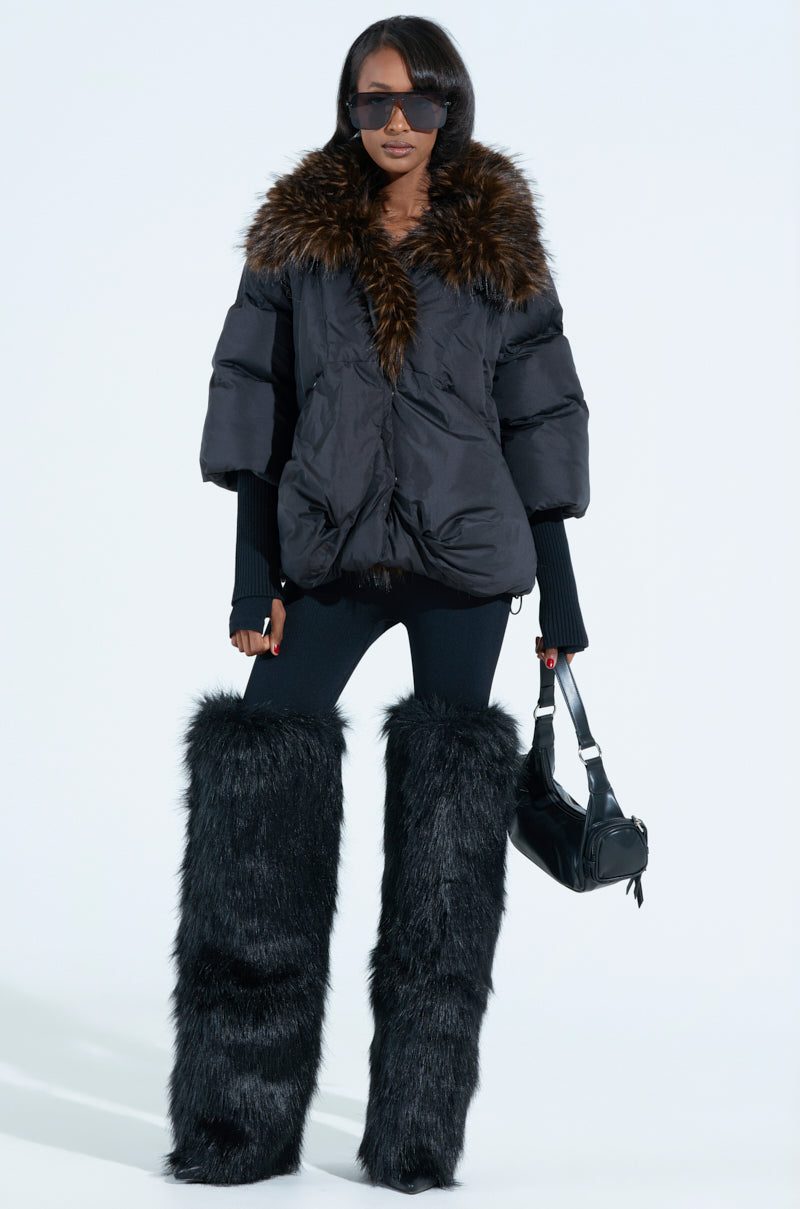 Sporting the FIRESIDE FUR TRIM PUFFER jacket, black leggings, and tall furry boots, the individual pairs a sleek black handbag with large dark sunglasses against a light background for an effortlessly stylish look.