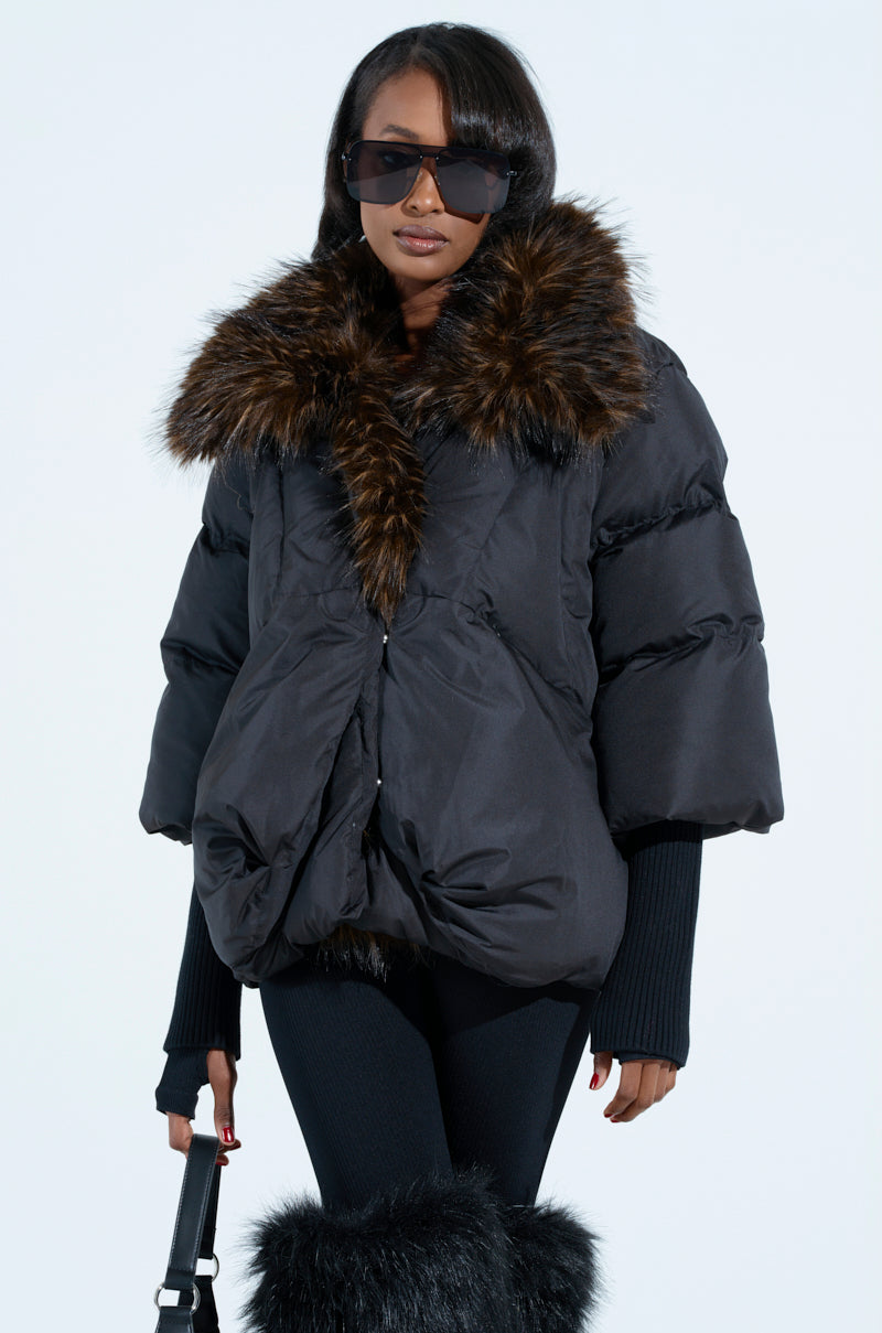 Dressed stylishly, the individual sports the FIRESIDE FUR TRIM PUFFER—a dark, oversized jacket with a plush faux fur collar. Paired with black leggings and boots, they have long hair and sunglasses while holding a black bag against a simple light background.