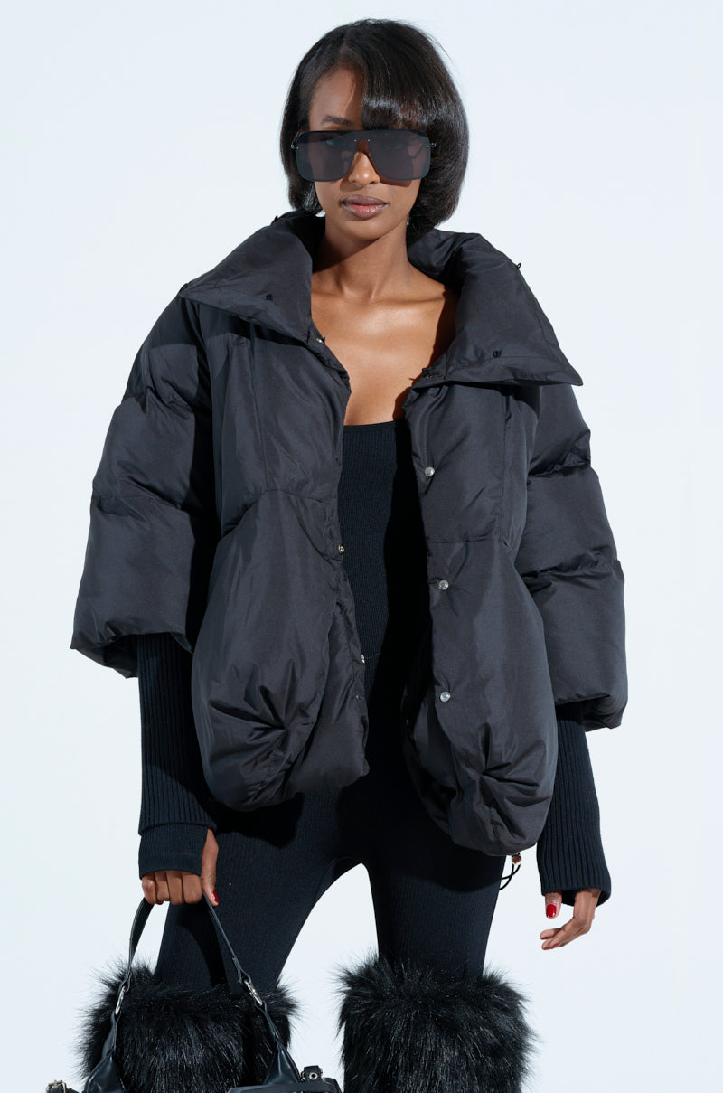 Against a light backdrop, someone models the FIRESIDE FUR TRIM PUFFER paired with oversized sunglasses. Sporting a chic bob and stylishly layered sleeves, they carry a black bag with fur accents and wear furry boots to complete the ensemble.