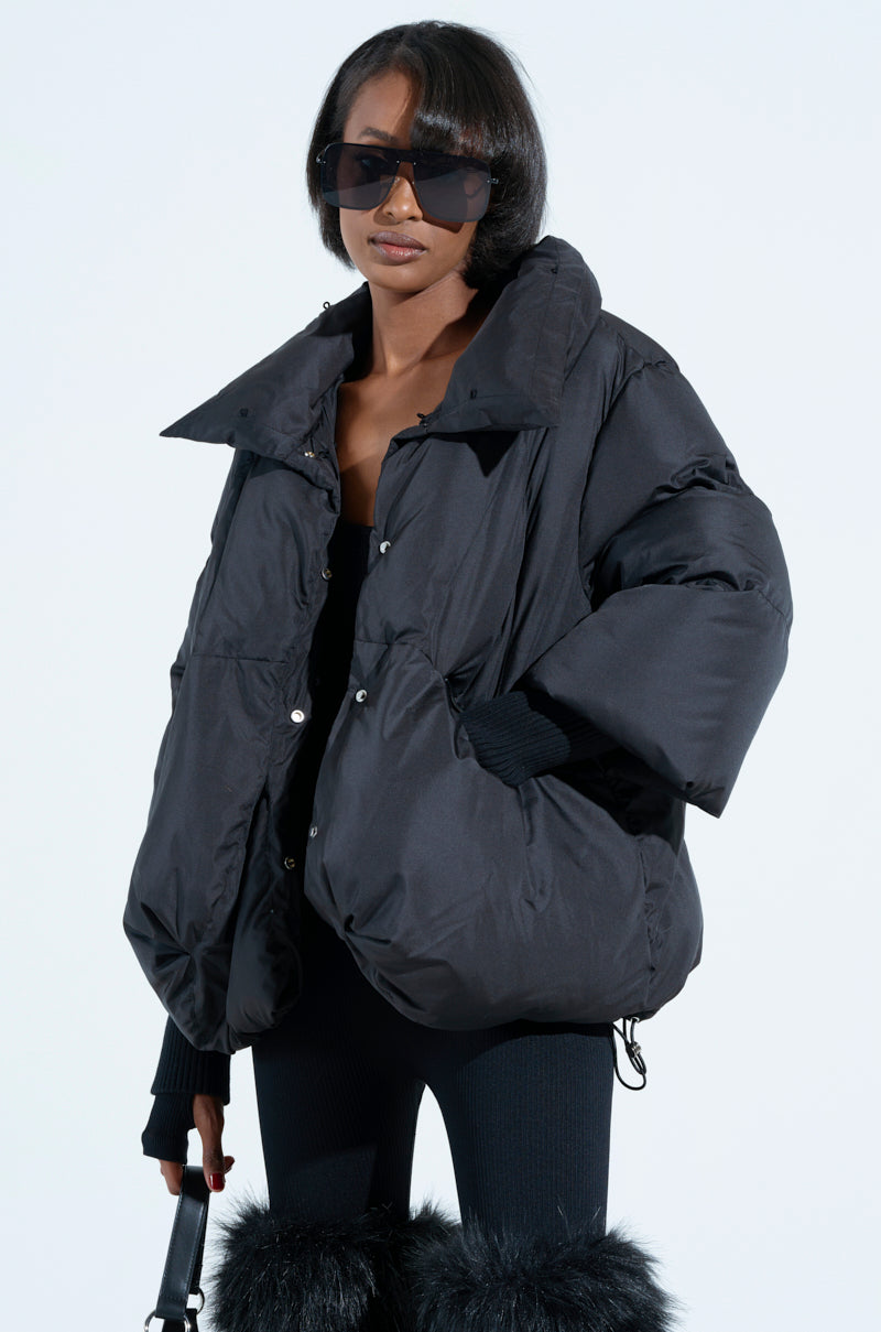 Against a light background, someone in a chic FIRESIDE FUR TRIM PUFFER, large sunglasses, black pants with fur details stands holding a black handbag.