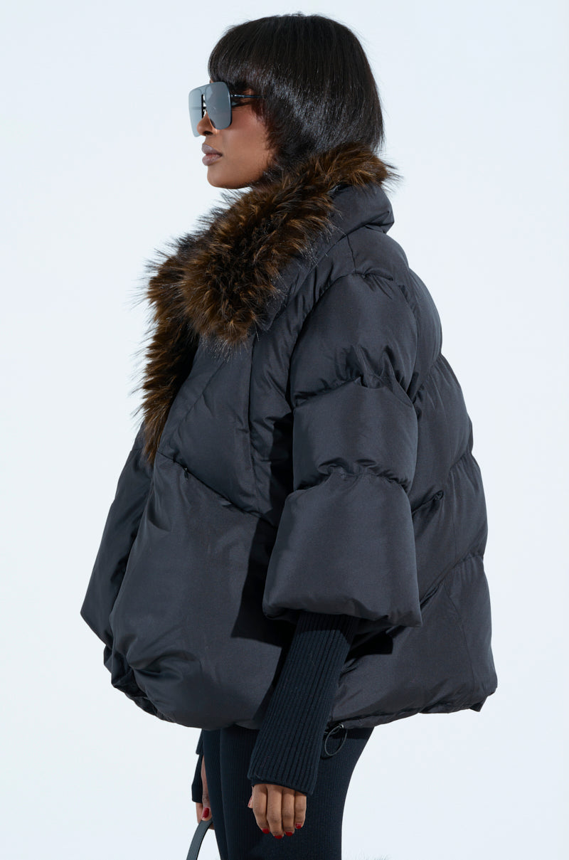 Stylishly facing left against a plain background, the person wears oversized sunglasses and the FIRESIDE FUR TRIM PUFFER. This chic black jacket features a fur-lined collar, wide sleeves, and a voluminous shape.