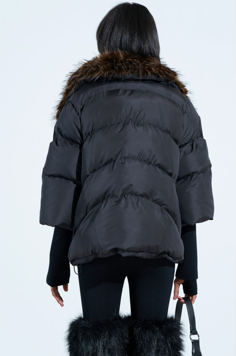 From behind, someone showcases a stylish look with long dark hair cascading down as they wear the FIRESIDE FUR TRIM PUFFER, featuring a black fur trim and brown fur-lined collar. They complete the outfit with black pants, knee-high plush faux fur boots, and a purse.