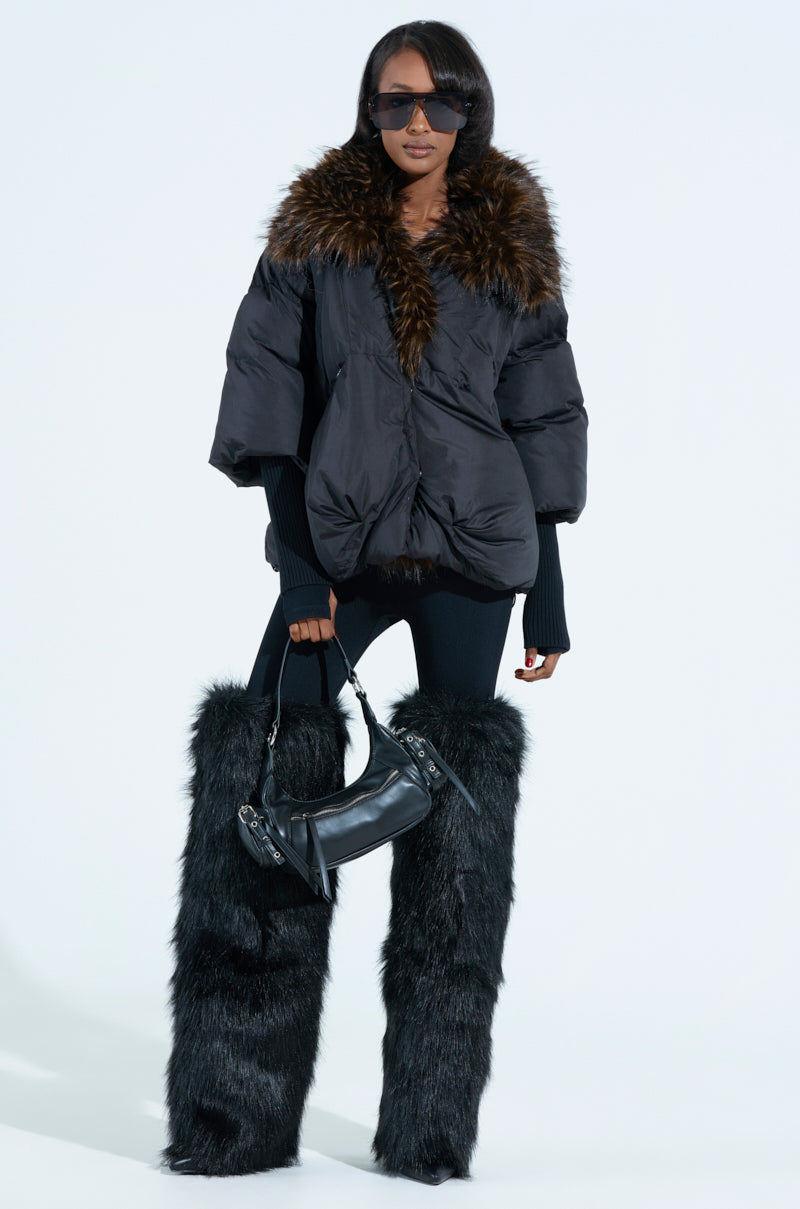 A woman showcases chic outerwear in a FIRESIDE FUR TRIM PUFFER with a plush faux fur collar, oversized black sunglasses, and sleek fur boots. Elegantly posing with her stylish handbag against a plain backdrop, she achieves an effortlessly glamorous look.