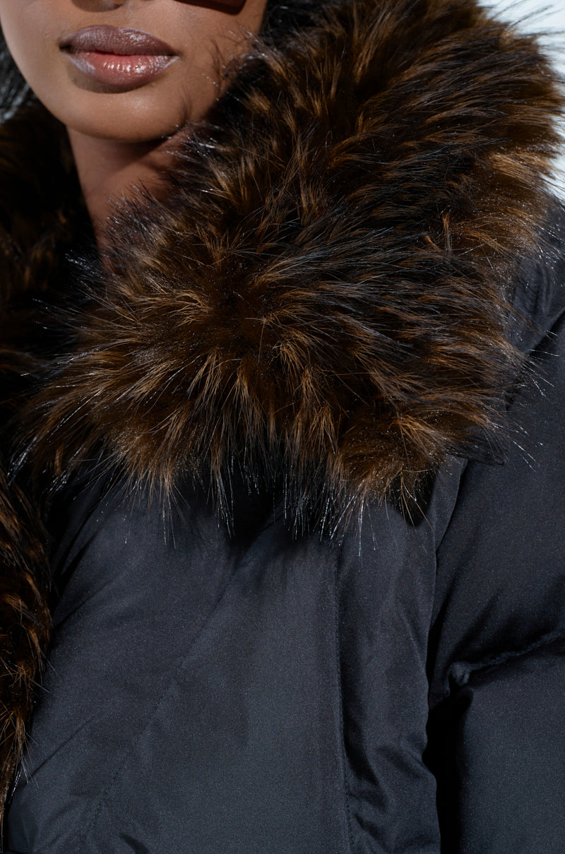 A person in chic outerwear wears the FIRESIDE FUR TRIM PUFFER in black, showcasing the plush faux fur collar. The close-up image highlights the coat's texture and fur trim, with a glimpse of their lower face slightly visible.
