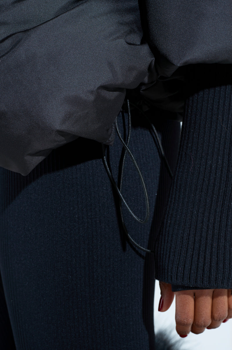 A close-up shows a person in chic FIRESIDE FUR TRIM PUFFER outerwear, featuring a dark, puffy jacket with ribbed knit sleeves. One hand is partially visible with relaxed fingers, while the blurred background accentuates the plush textures and subtle fur trim.