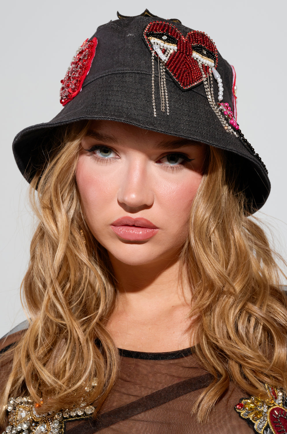 A person with long, wavy hair wears the WITH LOVE PATCH BUCKET HAT, which features beaded designs and embroidered patches, including a prominent face motif. They have a serious expression and are dressed in a sheer, embellished top.