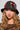 A person with long, wavy hair wears the WITH LOVE PATCH BUCKET HAT, which features beaded designs and embroidered patches, including a prominent face motif. They have a serious expression and are dressed in a sheer, embellished top.