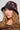 A person with long, wavy blonde hair wears the WITH LOVE PATCH BUCKET HAT, which is embellished with intricate red and white bead designs. They have a focused expression and are dressed in a black top decorated with colorful embellishments.