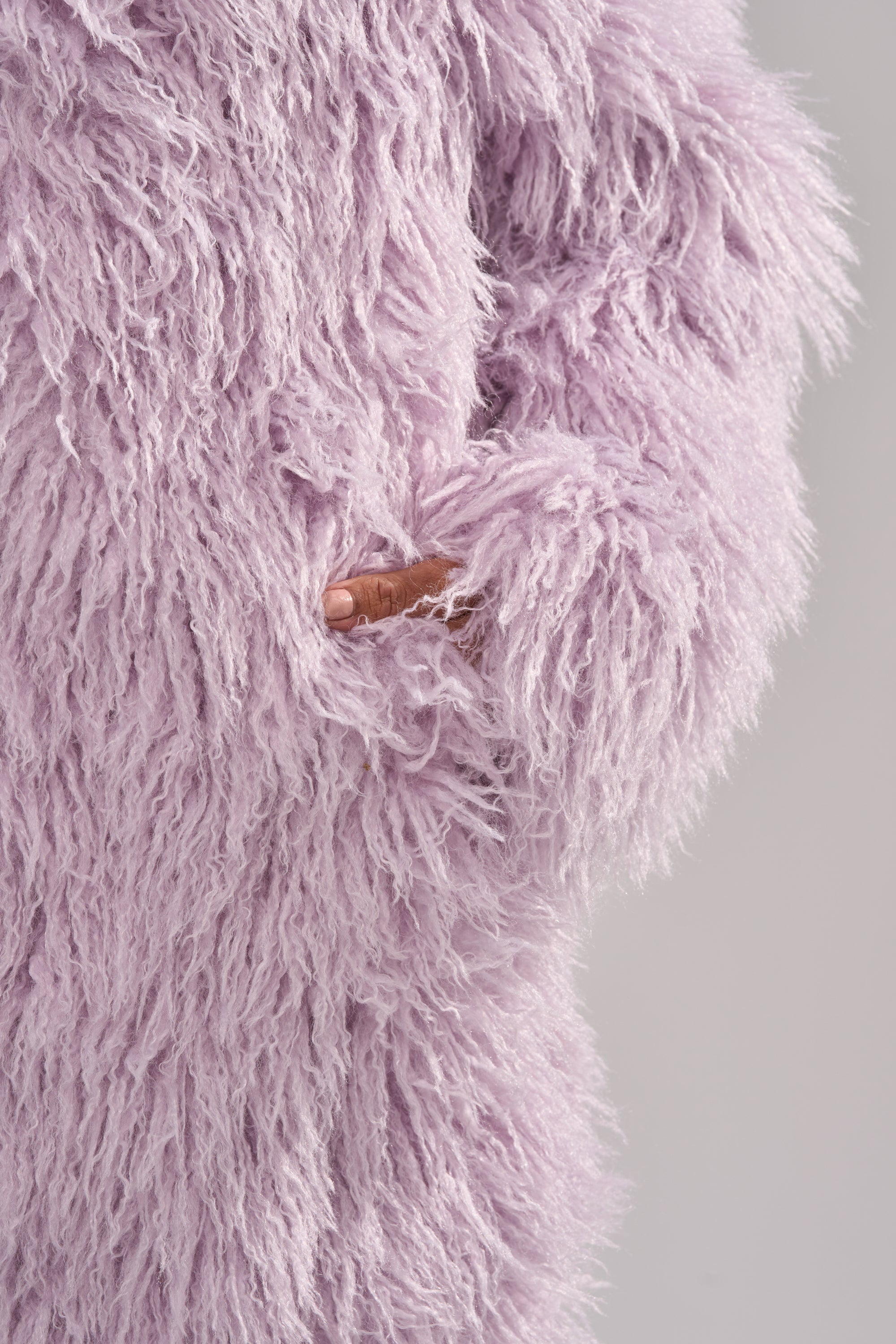 A hand is partly tucked into the pocket of the VIOLET SKIES FAUX FUR COAT, featuring a fluffy, lavender shaggy texture, set against a plain background. 9