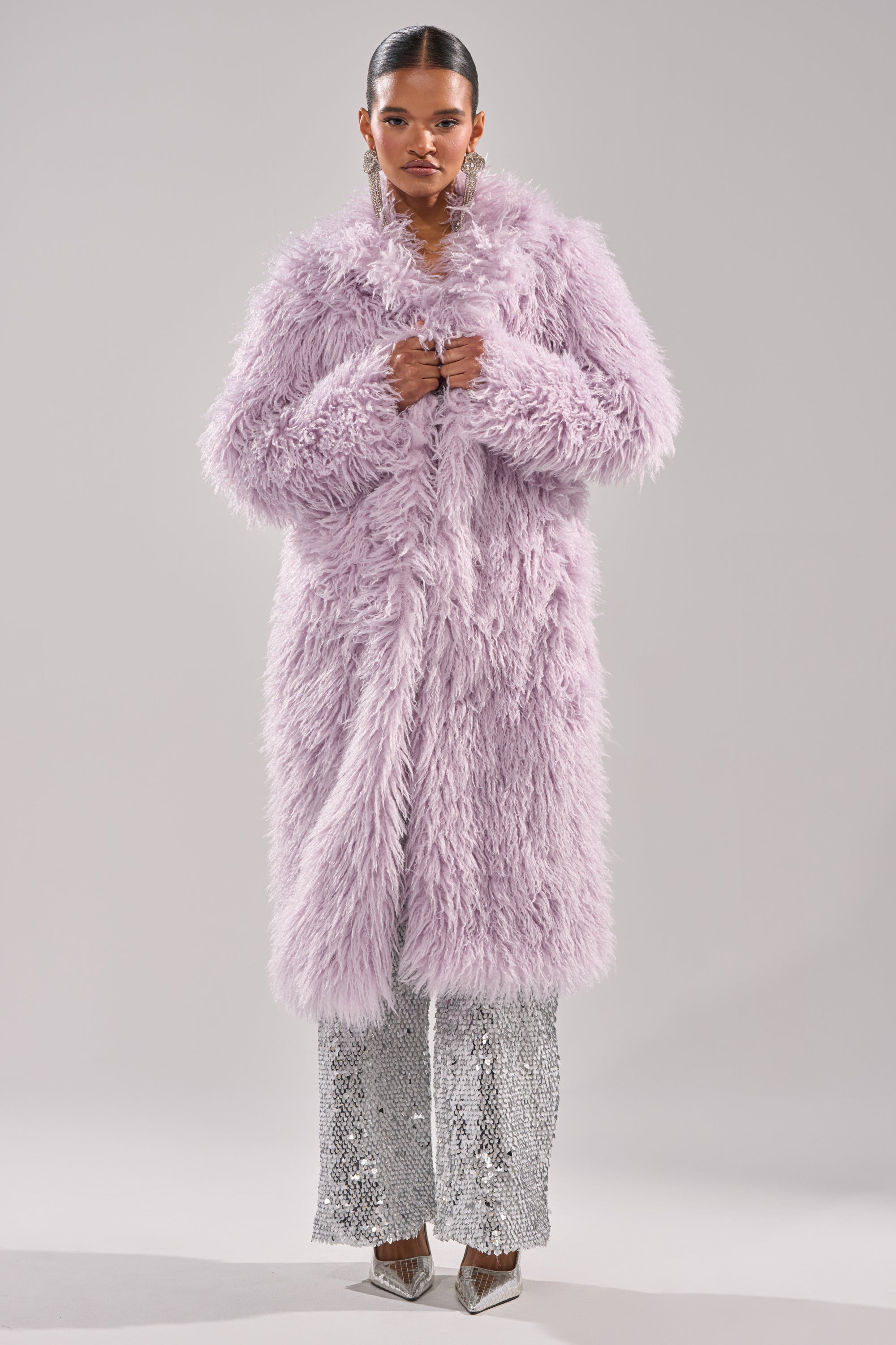 A woman faces forward in the AKIRA Label VIOLET SKIES FAUX FUR COAT, paired with silver sequin pants and pointed shoes. She wears her hair slicked back and large dangling earrings, set against a plain light background. 1