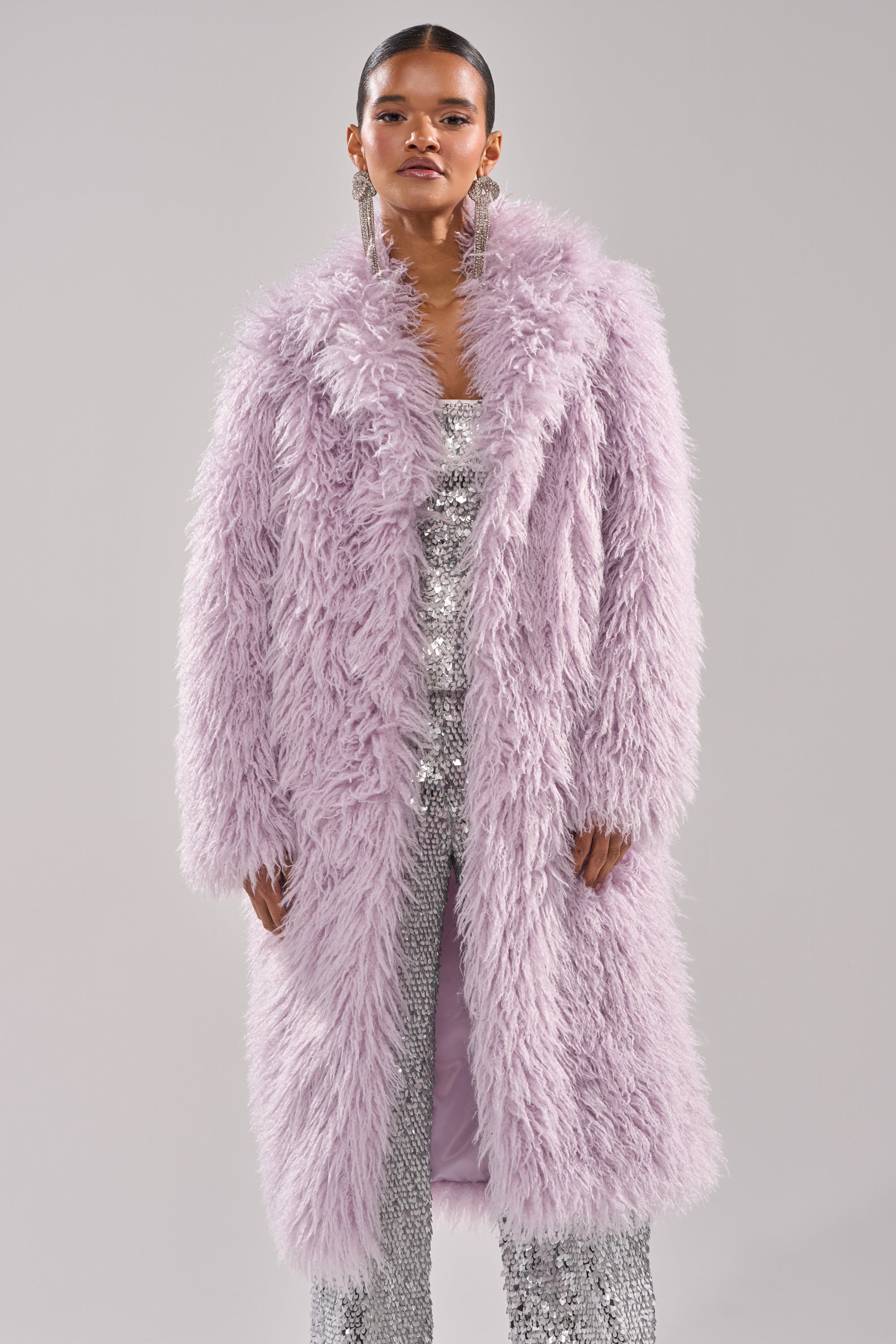 A woman faces forward against a light gray background, wearing the VIOLET SKIES FAUX FUR COAT over a sparkling silver sequin outfit, complemented by dangling earrings and slicked-back hair. 2
