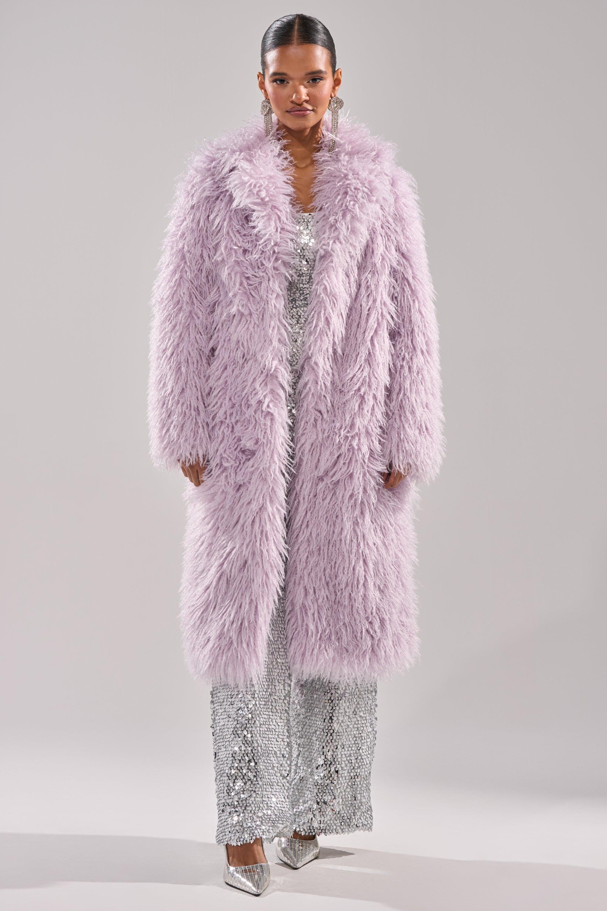 A woman faces forward in the VIOLET SKIES FAUX FUR COAT, styled over a sparkling silver sequin jumpsuit and metallic pointed heels, set against a plain light gray background. 6