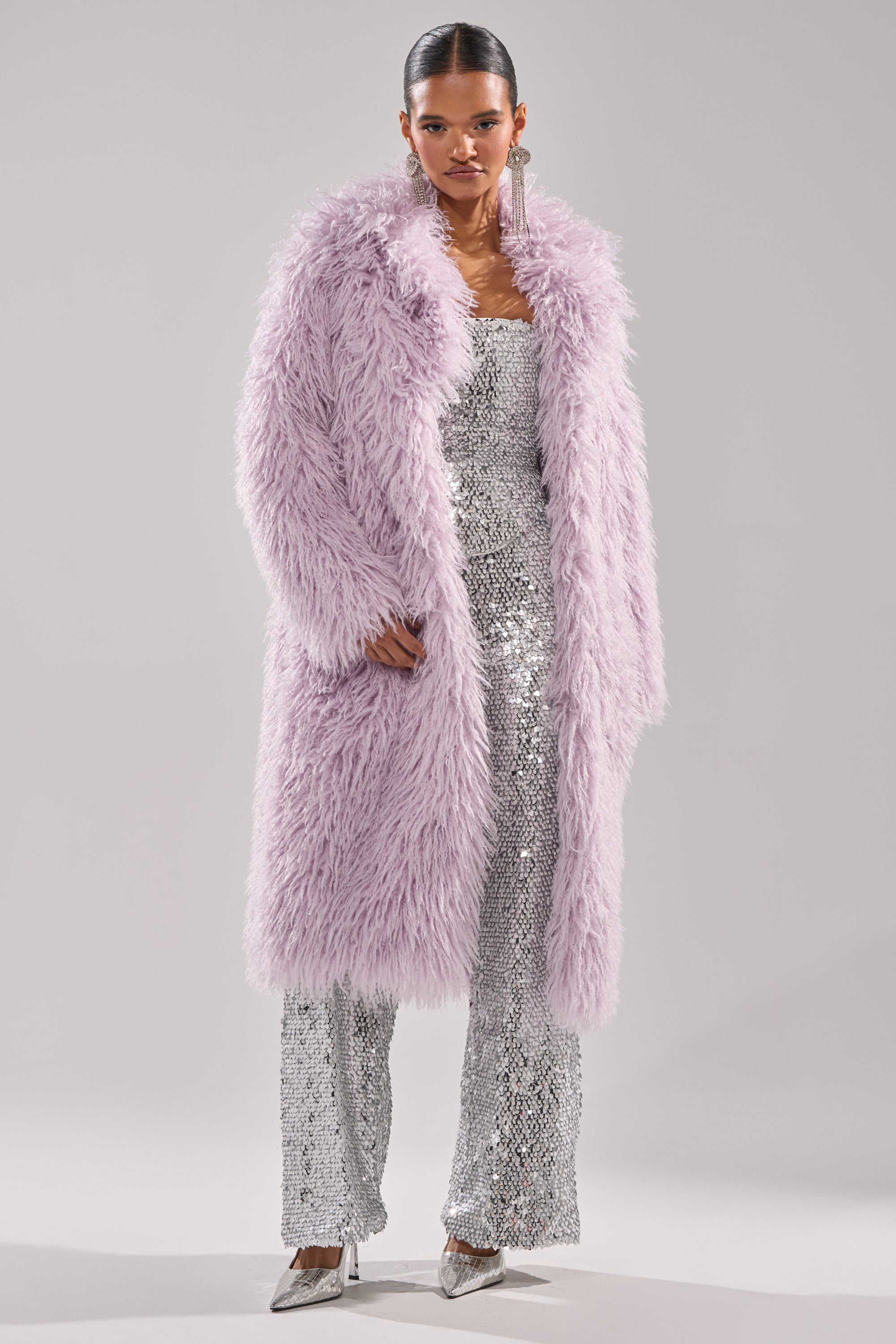 A woman wears the VIOLET SKIES FAUX FUR COAT in lavender over a shimmering silver sequin jumpsuit and silver heels. Her hair is sleek and pulled back, with large dangling earrings. The background is plain light gray. 7