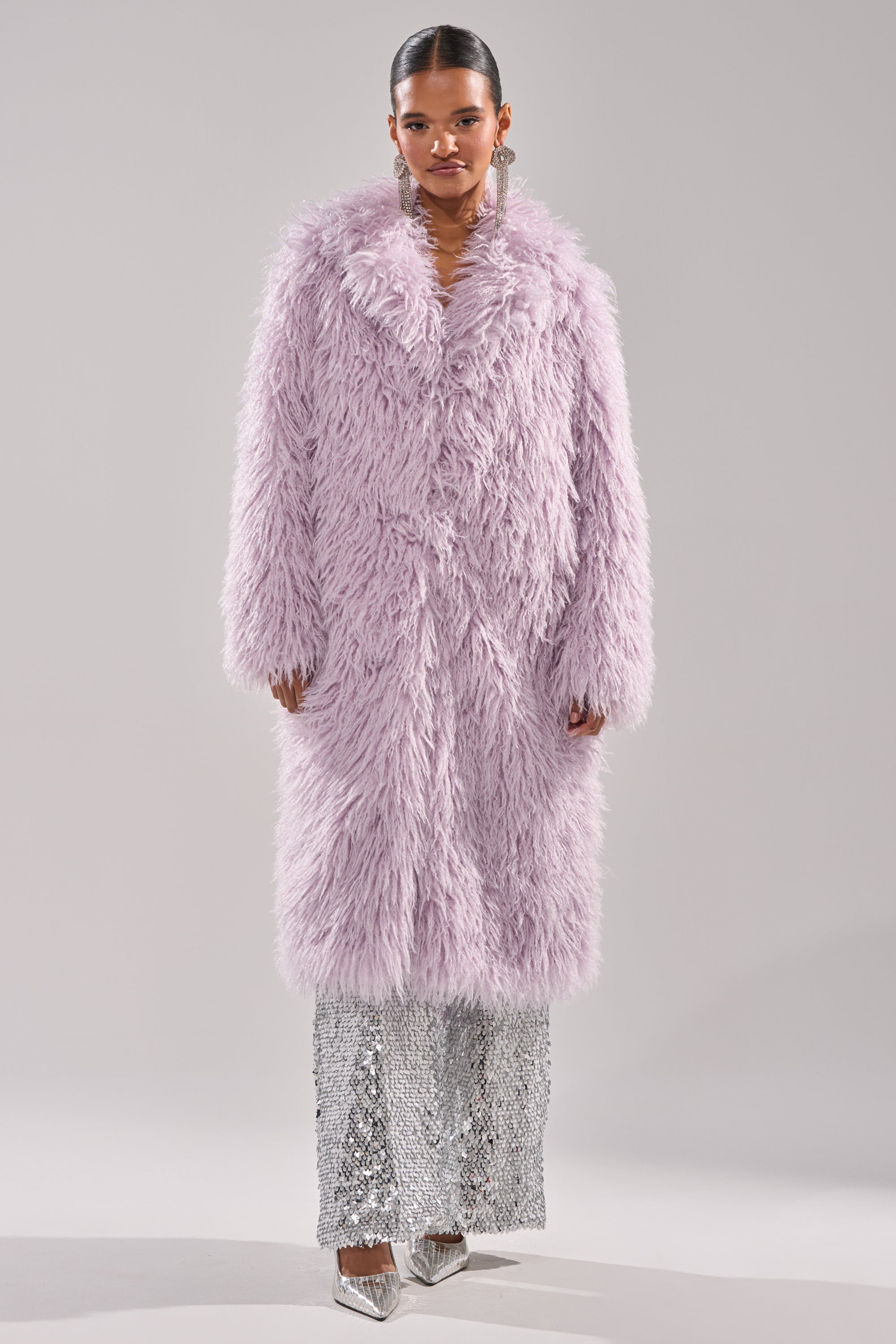 A woman faces forward in the VIOLET SKIES FAUX FUR COAT, styled over a silver sequined skirt and shiny silver heels. Her slicked-back hair and large dangling earrings complete the chic ensemble. 8