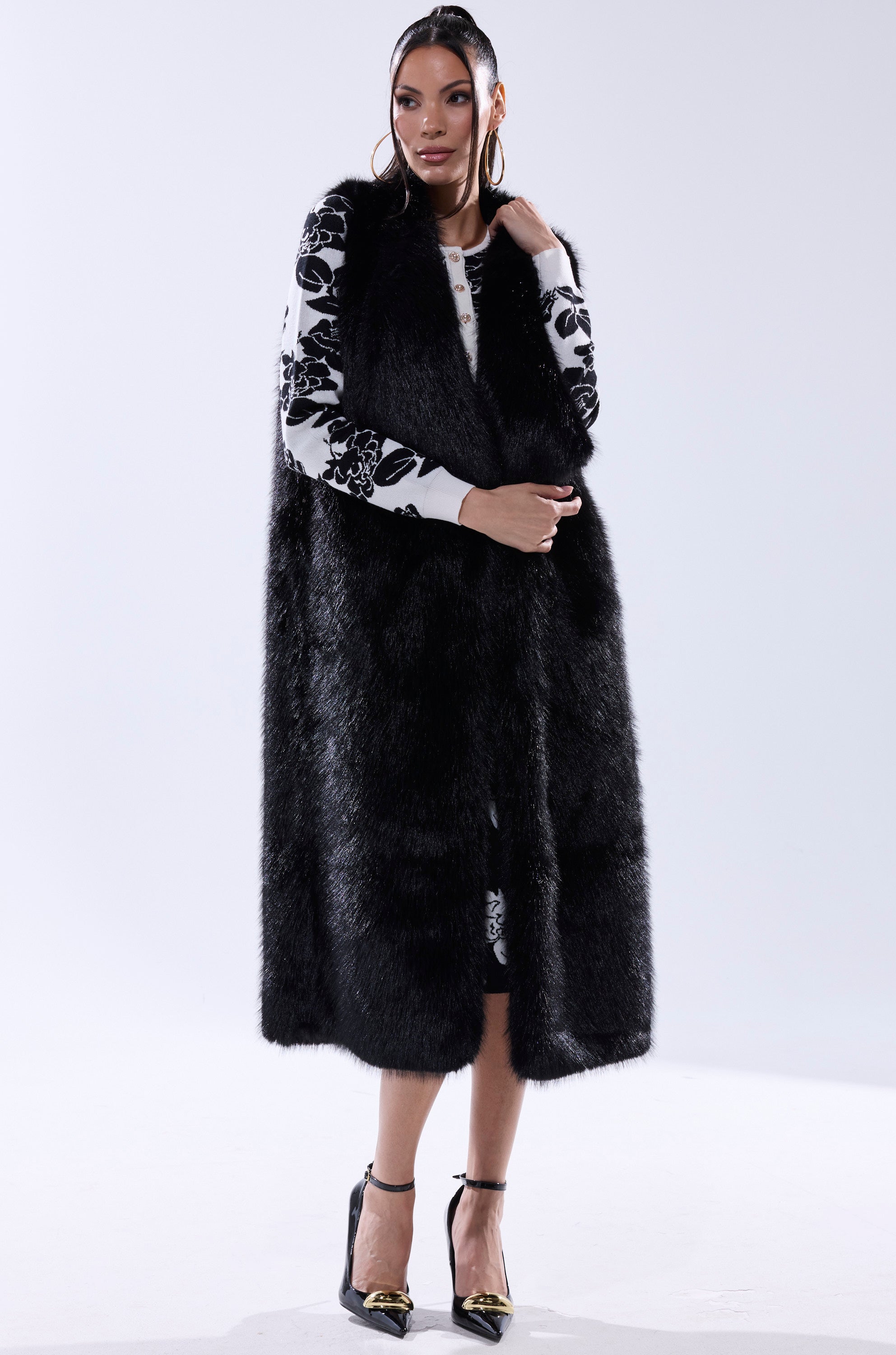 A woman styles the DOWNTIME FAUX FUR VEST IN BLACK over a white dress with black floral patterns, paired with hoop earrings and black strappy heels with gold tips. Her hair is in a sleek middle part.