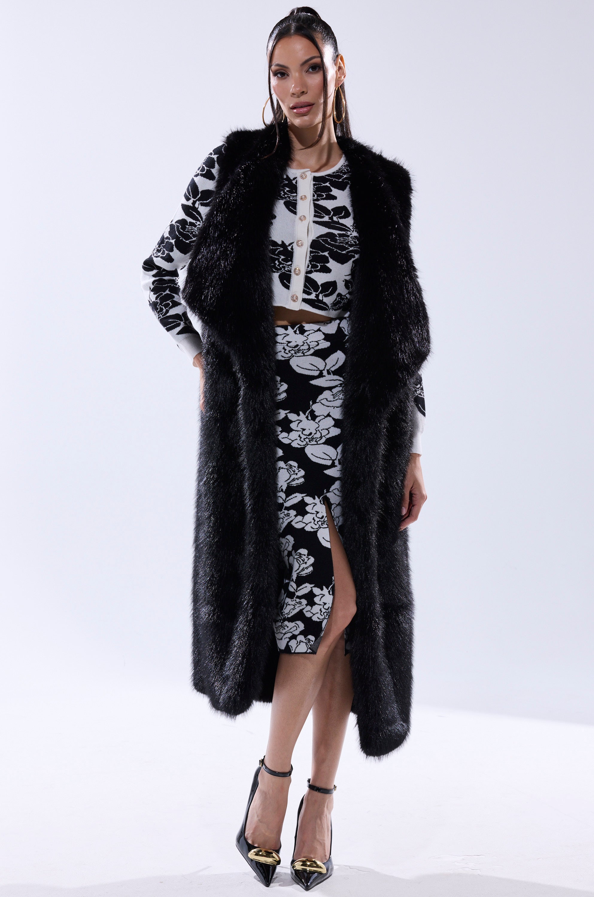 A woman wears the DOWNTIME FAUX FUR VEST IN BLACK over a black and white floral crop top and matching skirt with a front slit, styled with straight dark hair, hoop earrings, and black pointed heels with gold accents.
