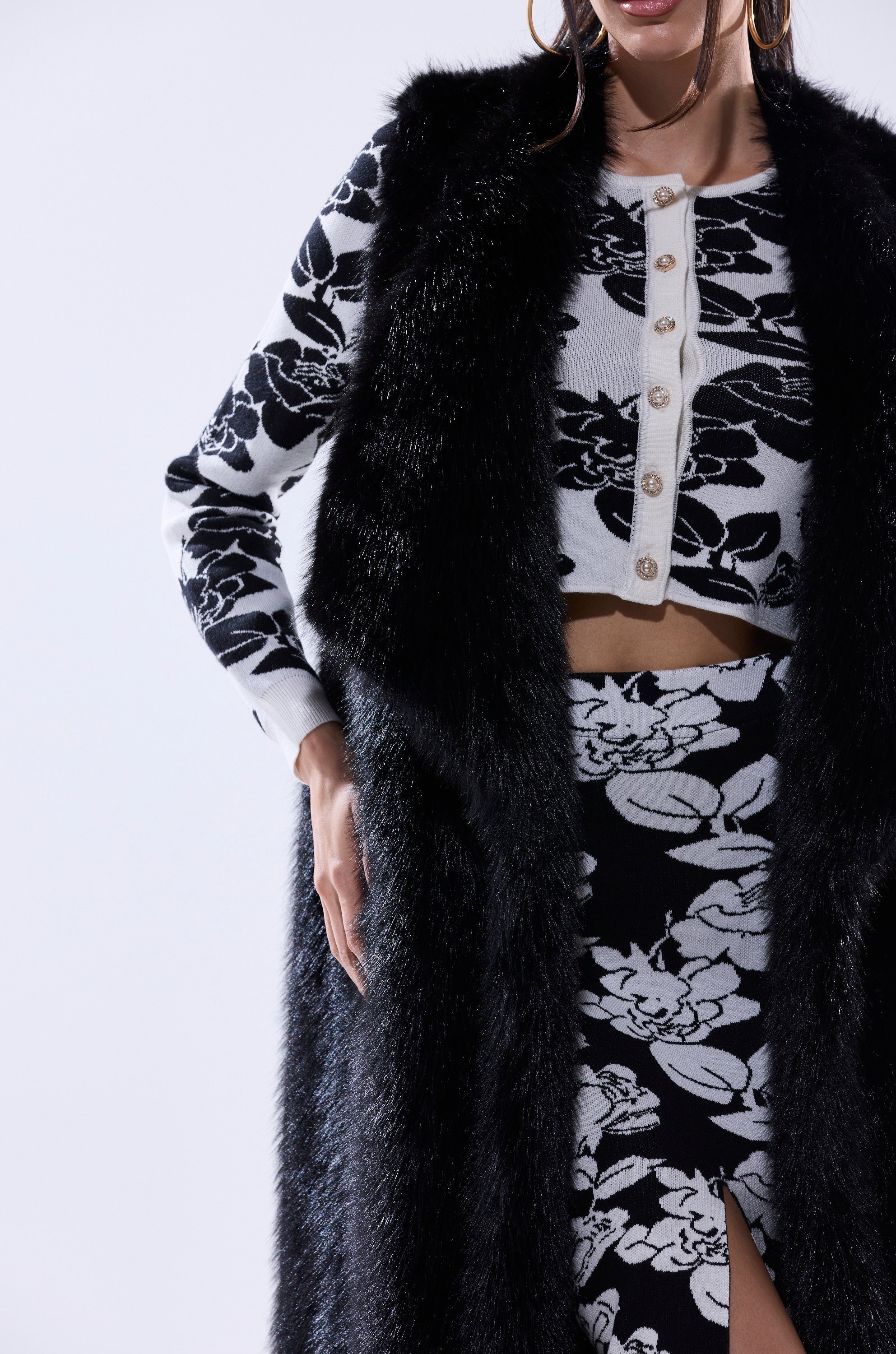 A woman wears the DOWNTIME FAUX FUR VEST IN BLACK over a white and black floral crop top with pearl buttons and a matching high-waisted floral skirt with a front slit. Her face is partially out of the frame.