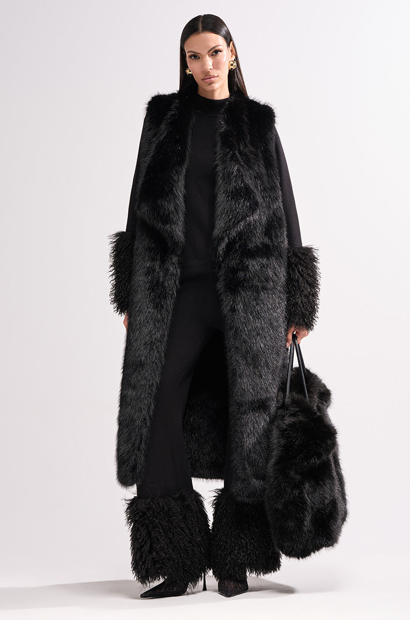 A woman with straight dark hair and a confident expression wears the DOWNTIME FAUX FUR VEST IN BLACK, paired with black pants featuring faux fur cuffs, and holds a large matching faux fur bag against a white background.