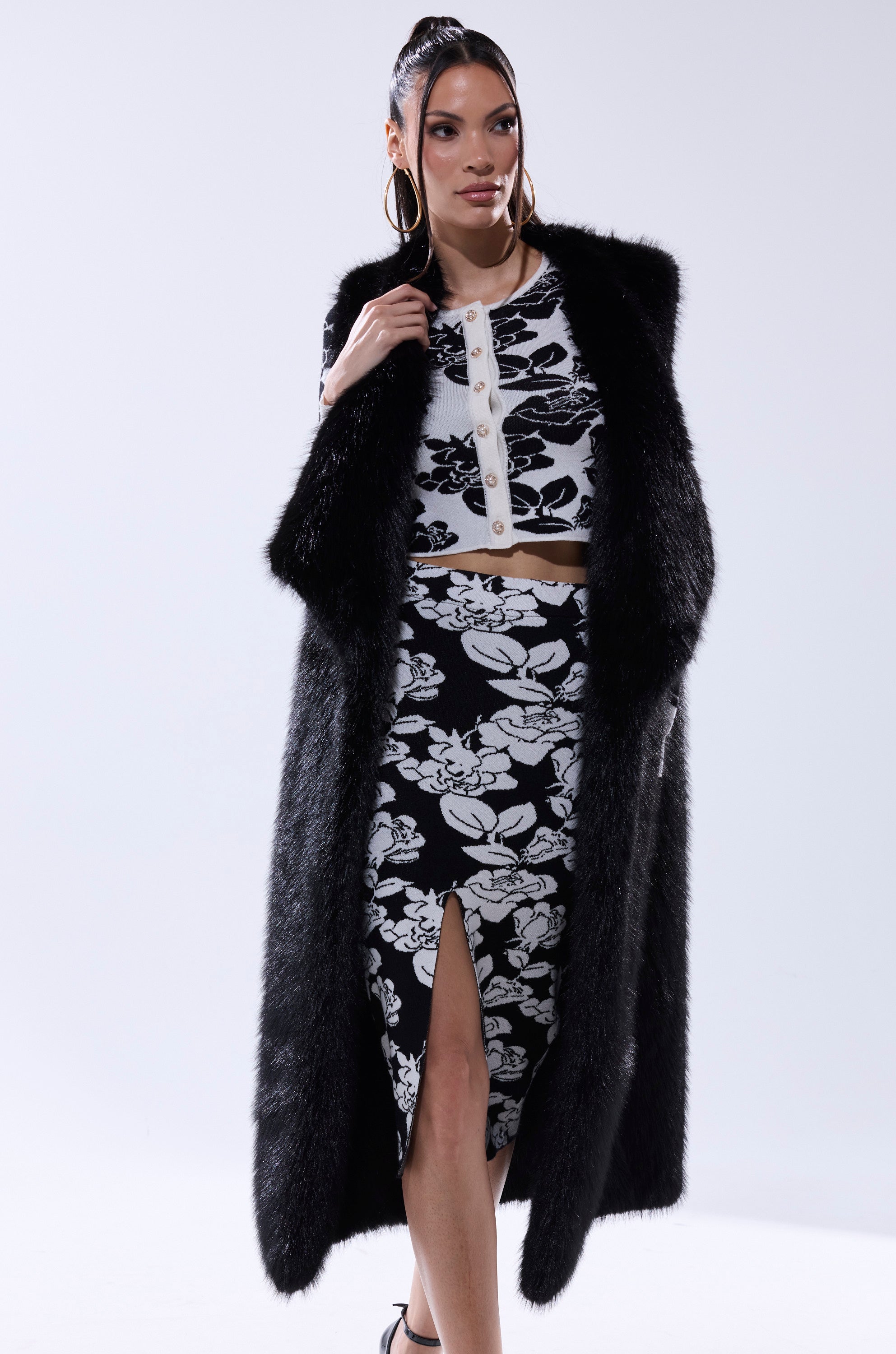 A woman poses in the DOWNTIME FAUX FUR VEST IN BLACK over a black and white floral crop top and matching front-slit skirt, accessorized with hoop earrings and a sleek ponytail.