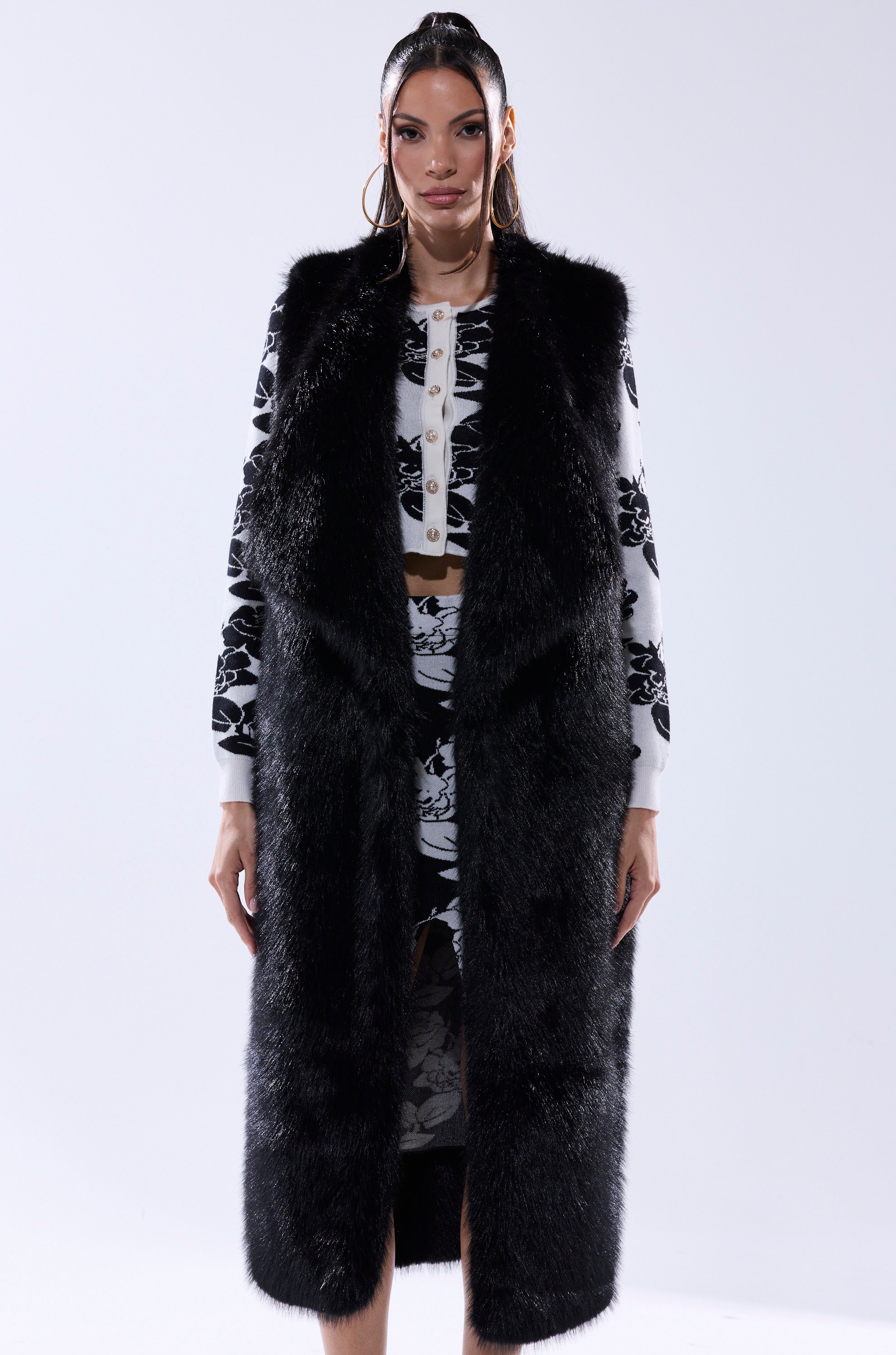 A woman with straight hair and hoop earrings stands confidently against a plain background, wearing the DOWNTIME FAUX FUR VEST IN BLACK over a black and white floral crop top and matching skirt.