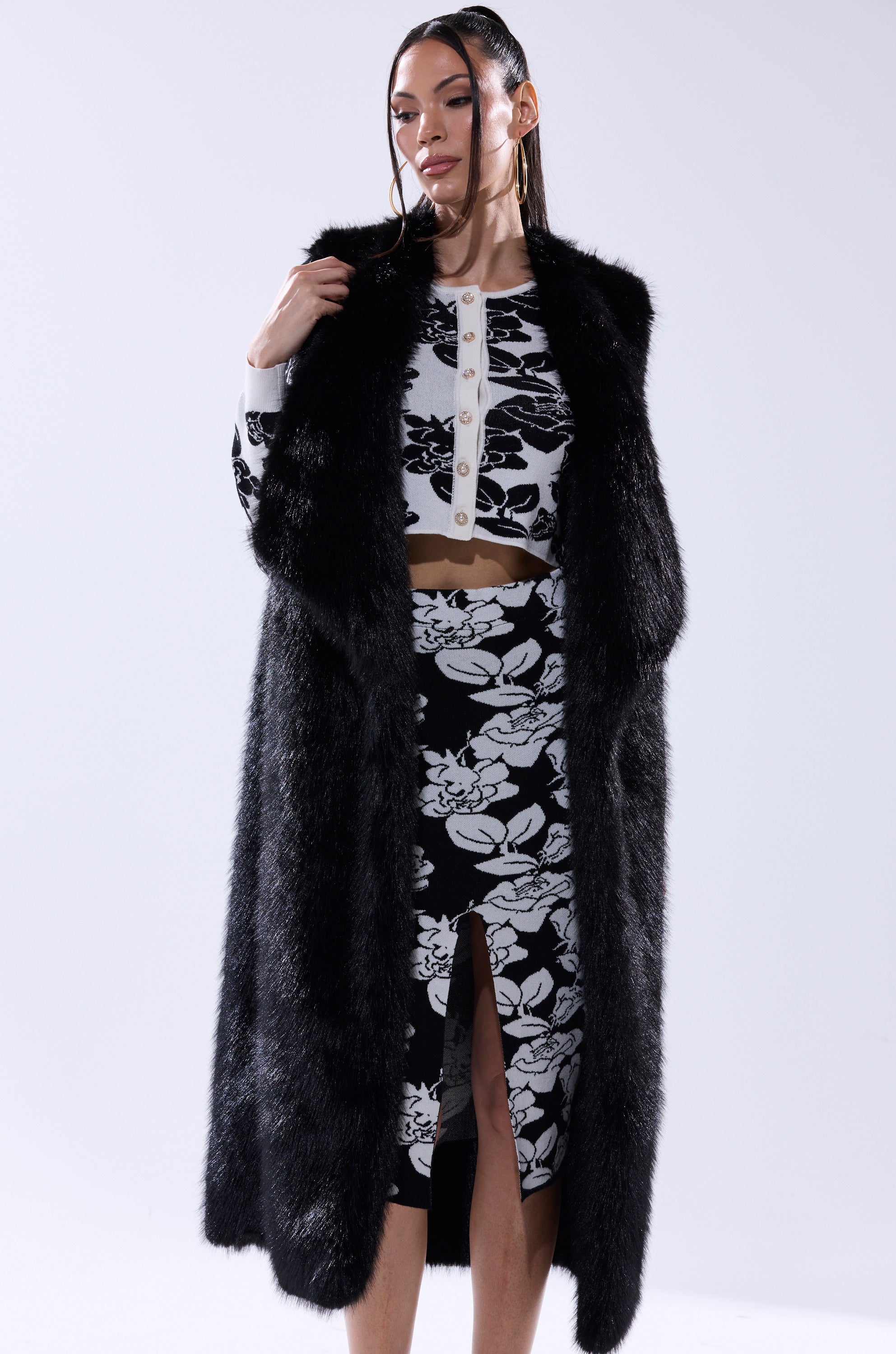 A woman wears the DOWNTIME FAUX FUR VEST IN BLACK over a white cropped floral cardigan and matching high-waisted skirt with a front slit, gazing to the side with one hand touching the vest.