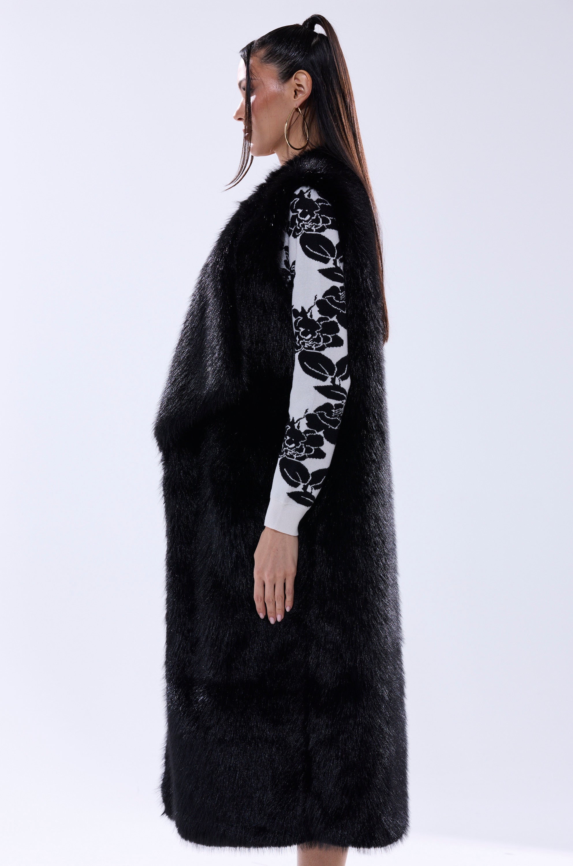 A woman with long, straight hair stands in profile against a plain light gray background, wearing the DOWNTIME FAUX FUR VEST IN BLACK over a white top with black floral patterns.