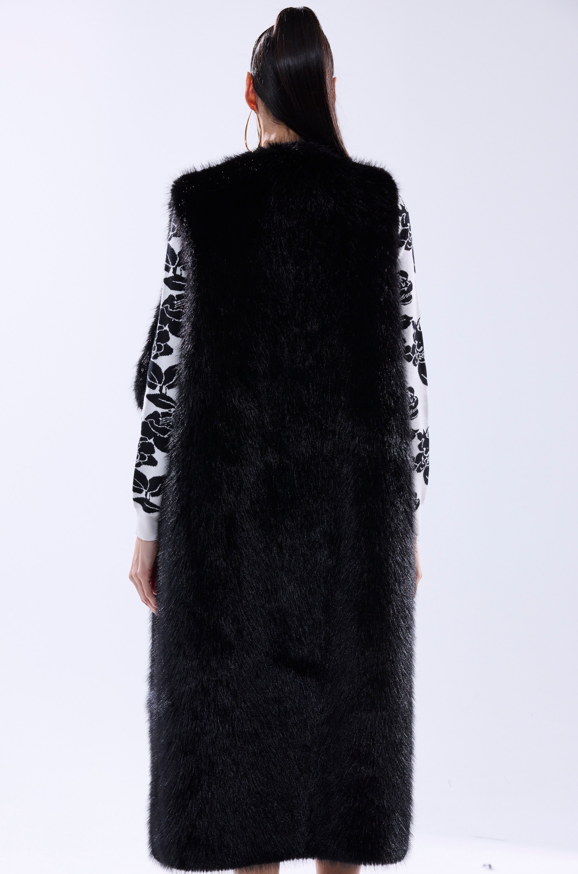 A person stands facing away, wearing the DOWNTIME FAUX FUR VEST IN BLACK—a longline silhouette—over a white top with black floral patterns. Their hair is styled in a high ponytail against a plain, light background.