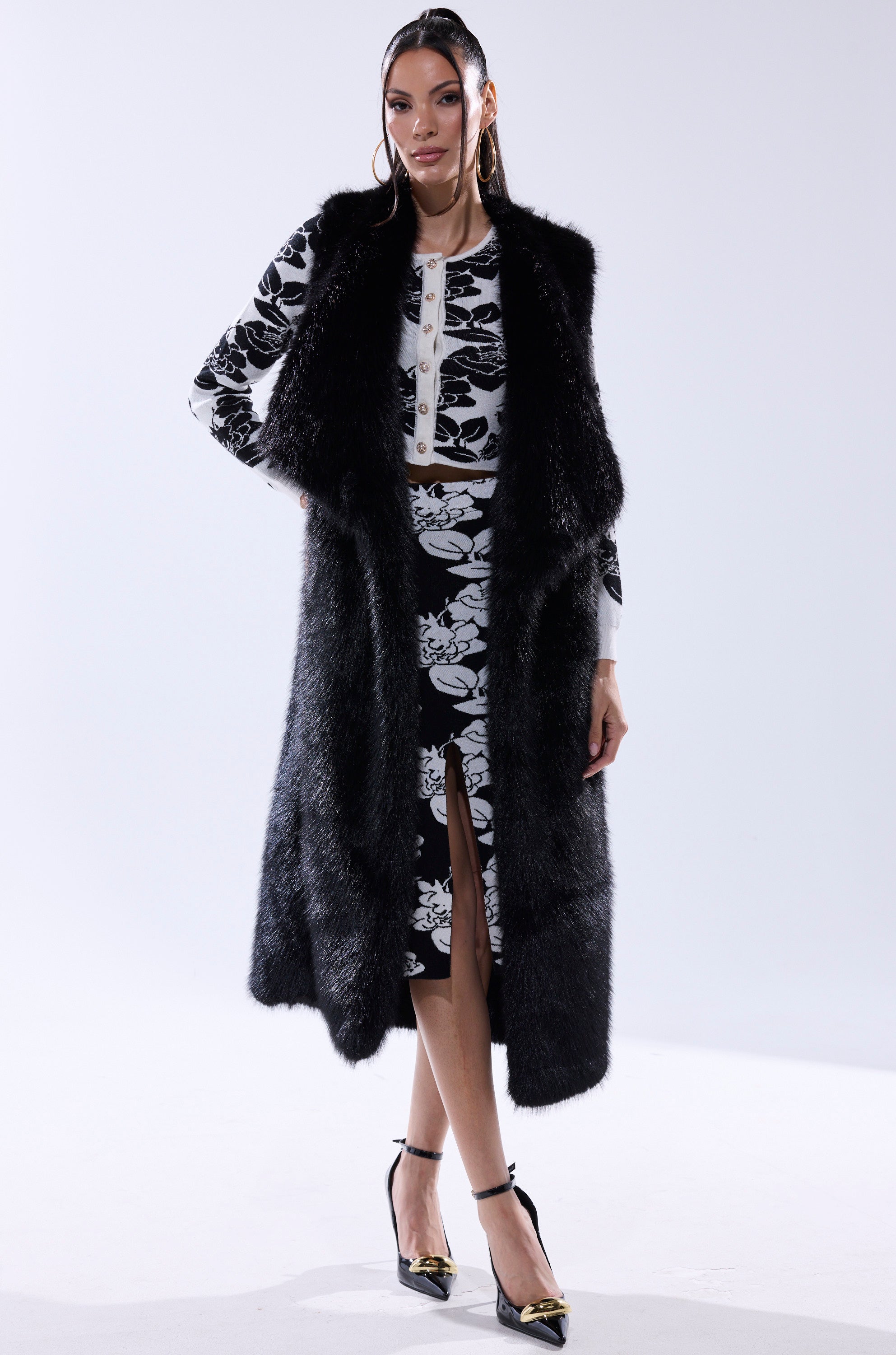 A woman stands against a white background wearing the DOWNTIME FAUX FUR VEST IN BLACK over a black and white floral print top and front-slit skirt, paired with black ankle-strap heels.