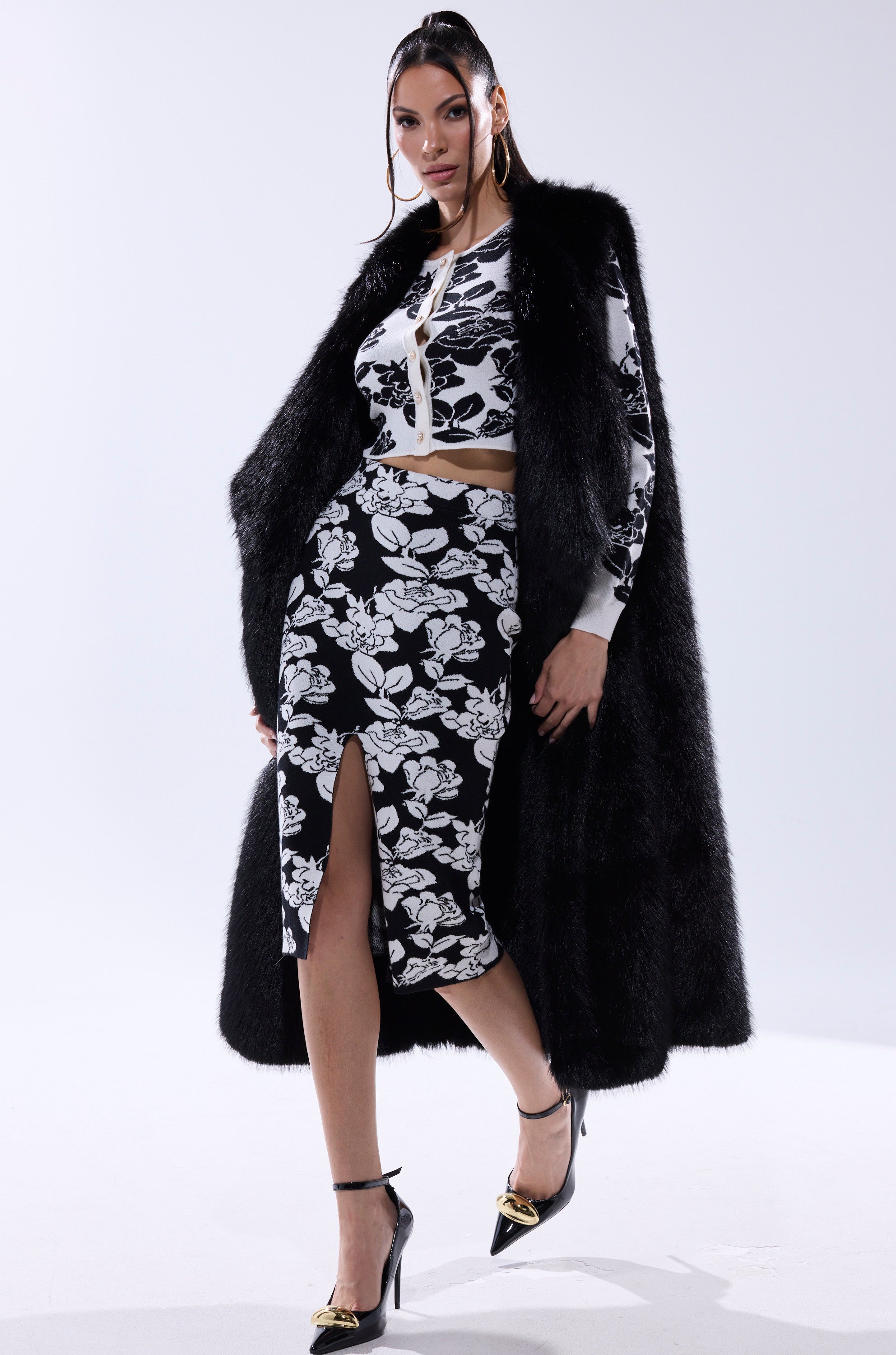 A woman models the DOWNTIME FAUX FUR VEST IN BLACK over a black and white floral crop top and skirt with a front slit, paired with gold-accented black heels and hoop earrings, against a plain white background.